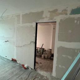 Leo Serwis Montaż i Demontaż - Wnętrze w trakcie remontu: ściany z płyt gipsowo-kartonowych, widoczne instalacje elektryczne, częściowo zaszpachlowane łączenia płyt, podłoga drewniana, w głębi pomieszczenia materiały budowlane.