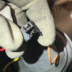 Marcin Fabicki - Dłoń w roboczej rękawicy trzyma czarną kostkę elektryczną z widocznymi stykami, obok widoczne przewody: pomarańczowy i żółty, oraz fragment reflektora samochodowego.