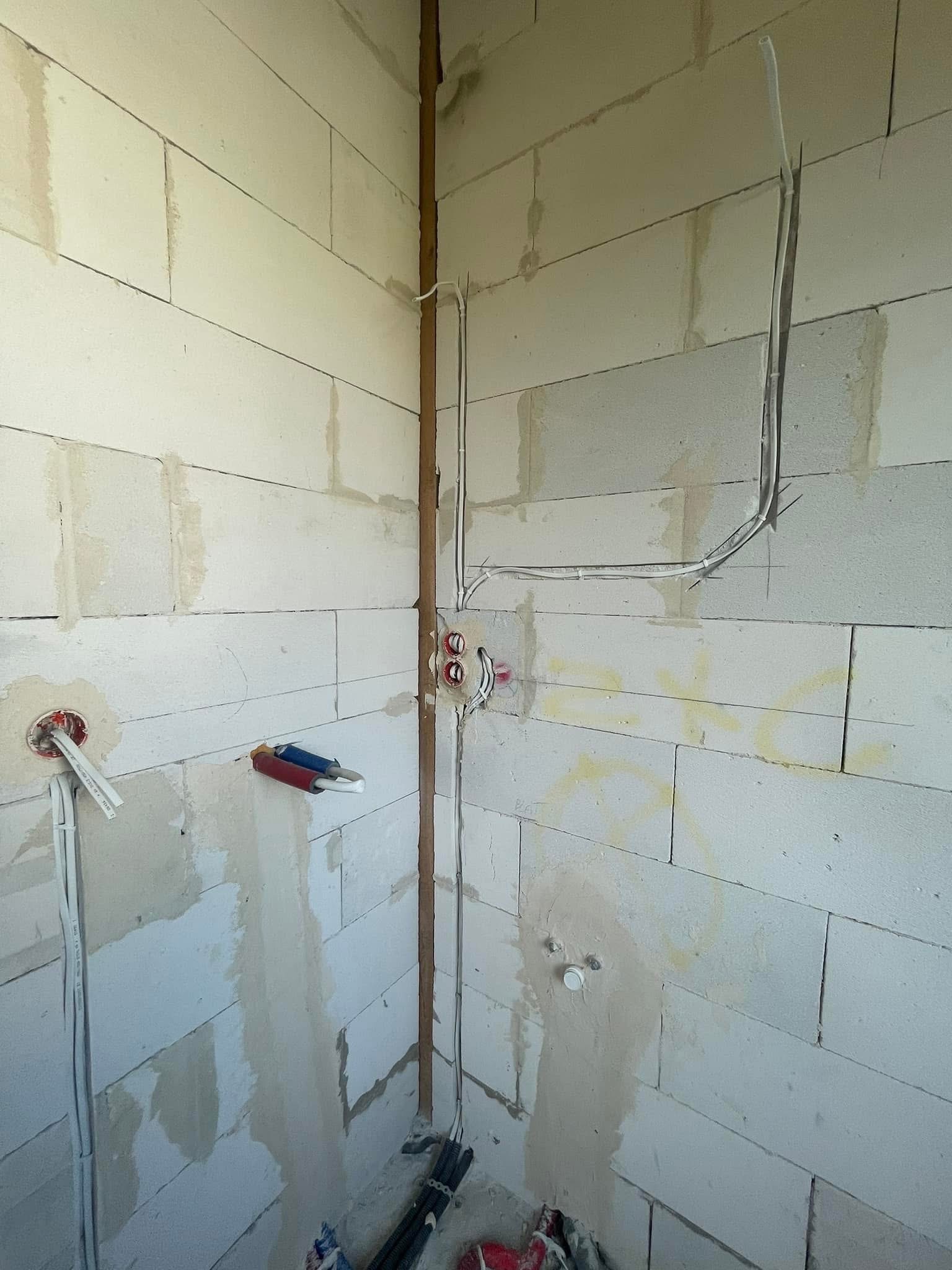 Surowy stan: Instalacja elektryczna w domu w trakcie budowy, widoczne przewody, puszki elektryczne i rury prowadzące kable na tle ściany z betonu komórkowego.