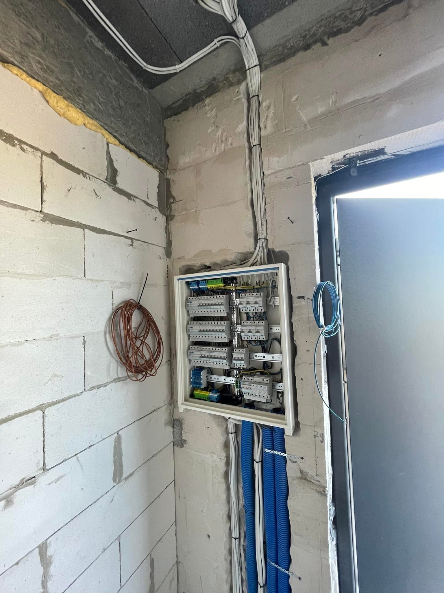 Rozdzielnia elektryczna w stanie deweloperskim, widoczne kable prowadzone po ścianie z bloczków, niebieskie rury karbowane i zwisający kabel przy ościeżnicy okna.