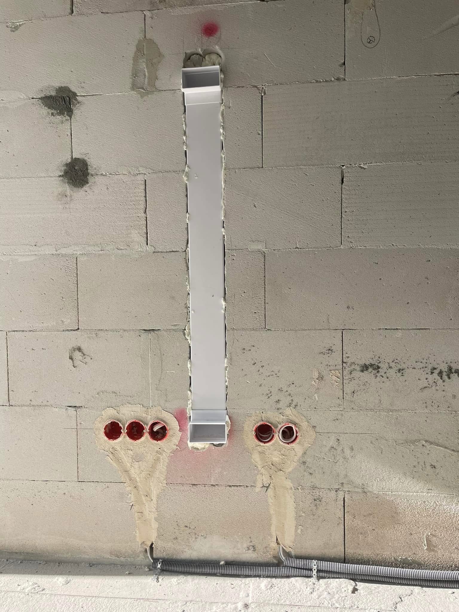 Surowa ściana z betonu komórkowego z zamontowaną, białą listwą maskującą pionowy przebieg instalacji elektrycznej oraz trzema czerwonymi puszkami podtynkowymi po lewej i dwiema po prawej stronie...