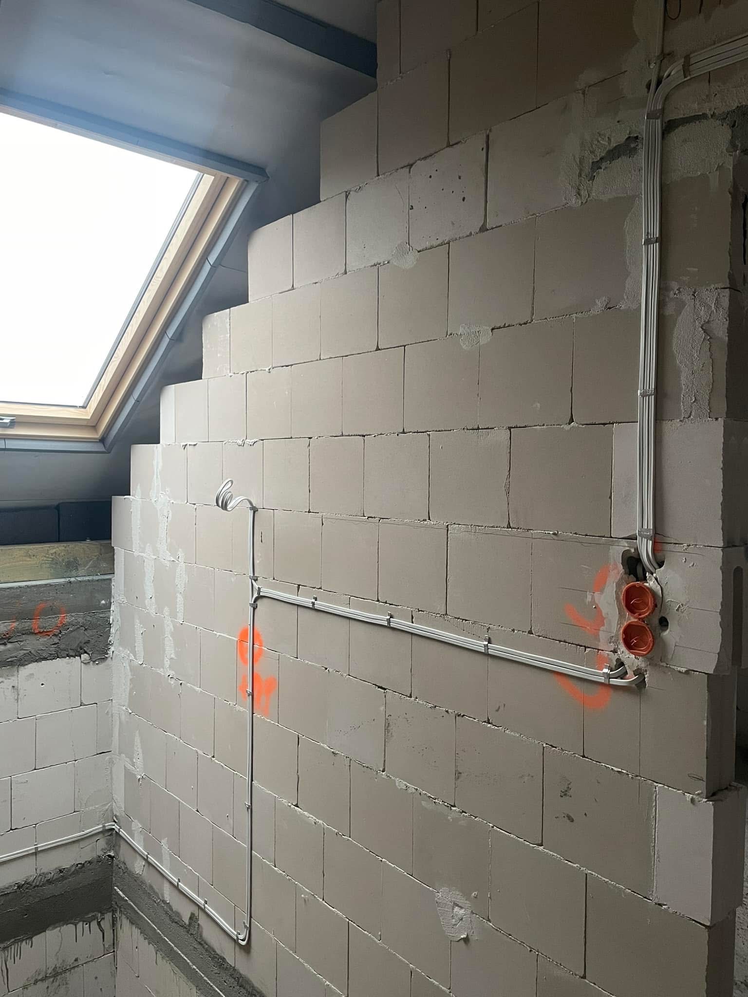 Surowa ściana z betonu komórkowego w stanie deweloperskim z zamontowanymi przewodami elektrycznymi w listwach i puszkami instalacyjnymi, oznaczona pomarańczową farbą, widoczne okno dachowe.