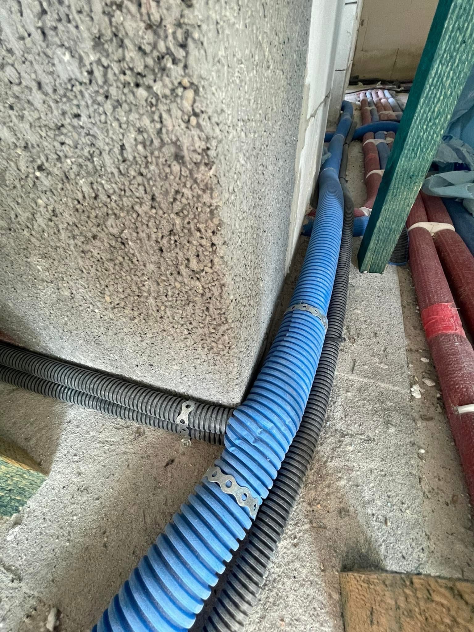 Rury instalacyjne w kolorze niebieskim, czarnym i czerwonym, prowadzone wzdłuż surowej ściany z betonu komórkowego w stanie deweloperskim, zabezpieczone metalowymi uchwytami.