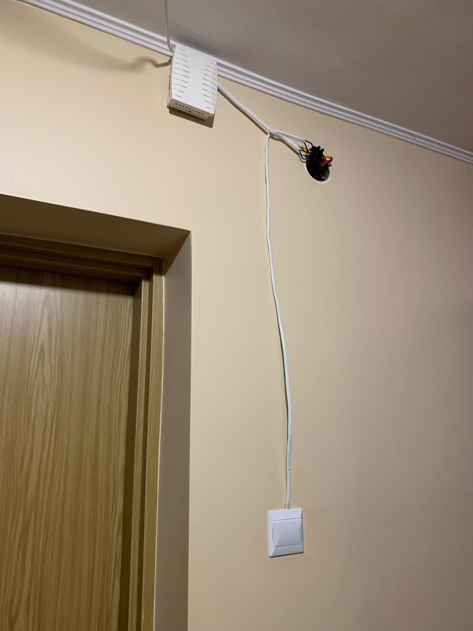 Biała ściana z widoczną instalacją elektryczną: router na górze, kabel prowadzący do gniazdka i otwór w suficie z wystającymi przewodami.