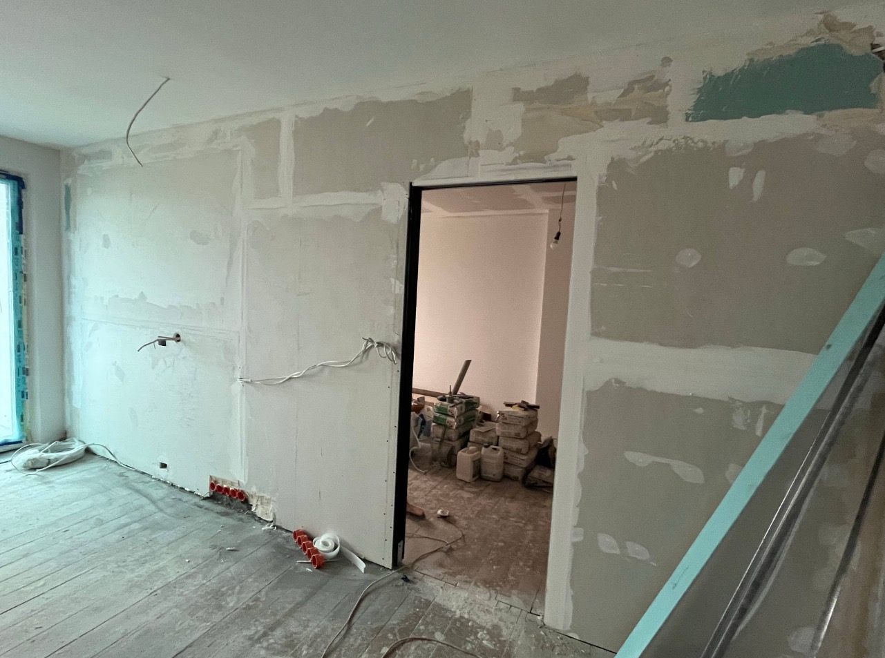 Wnętrze w trakcie remontu: ściany z płyt gipsowo-kartonowych, widoczne instalacje elektryczne, częściowo zaszpachlowane łączenia płyt, podłoga drewniana, w głębi pomieszczenia materiały budowlane.