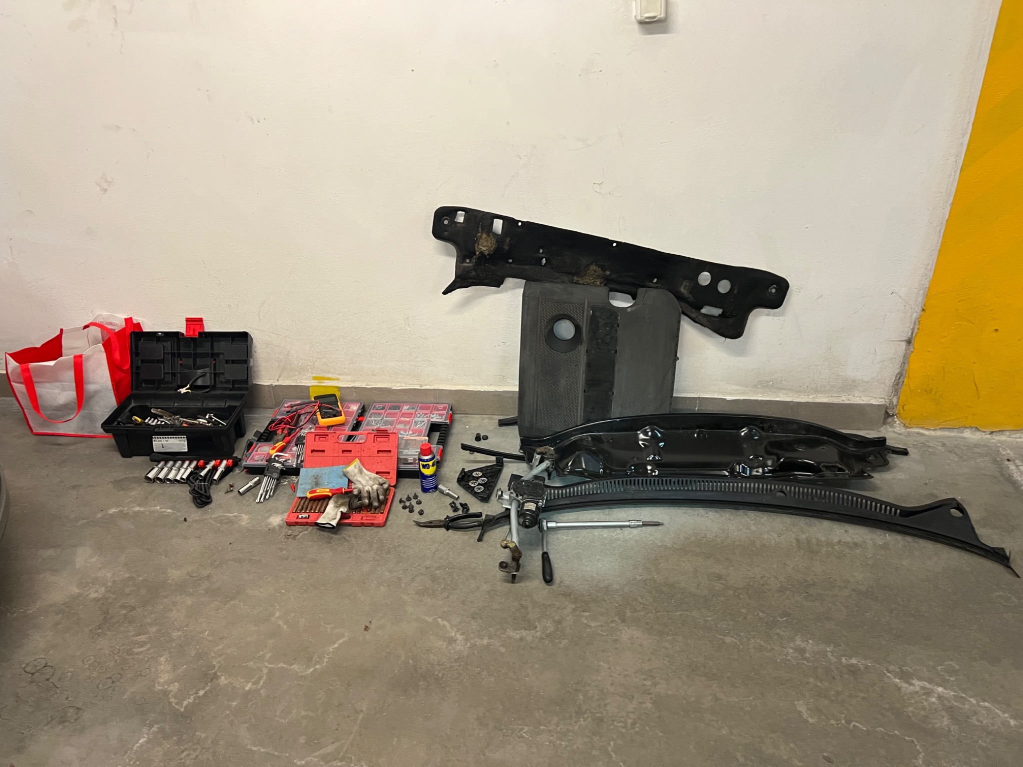 Rozłożone elementy podszybia samochodowego VW Polo obok skrzynki z narzędziami, kluczy nasadowych, multimetru i preparatu WD-40 na betonowej posadzce garażu z żółtym akcentem na ścianie.