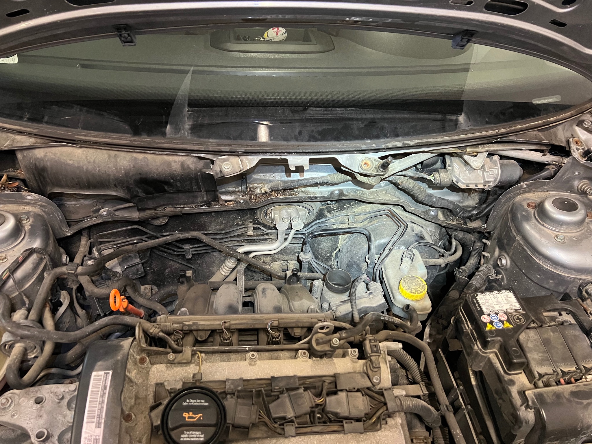 Widok pod maską samochodu VW Polo, z widocznym silnikiem, akumulatorem i elementami układu wycieraczek pod szybą. W tle deska rozdzielcza i mała piłka do rugby.