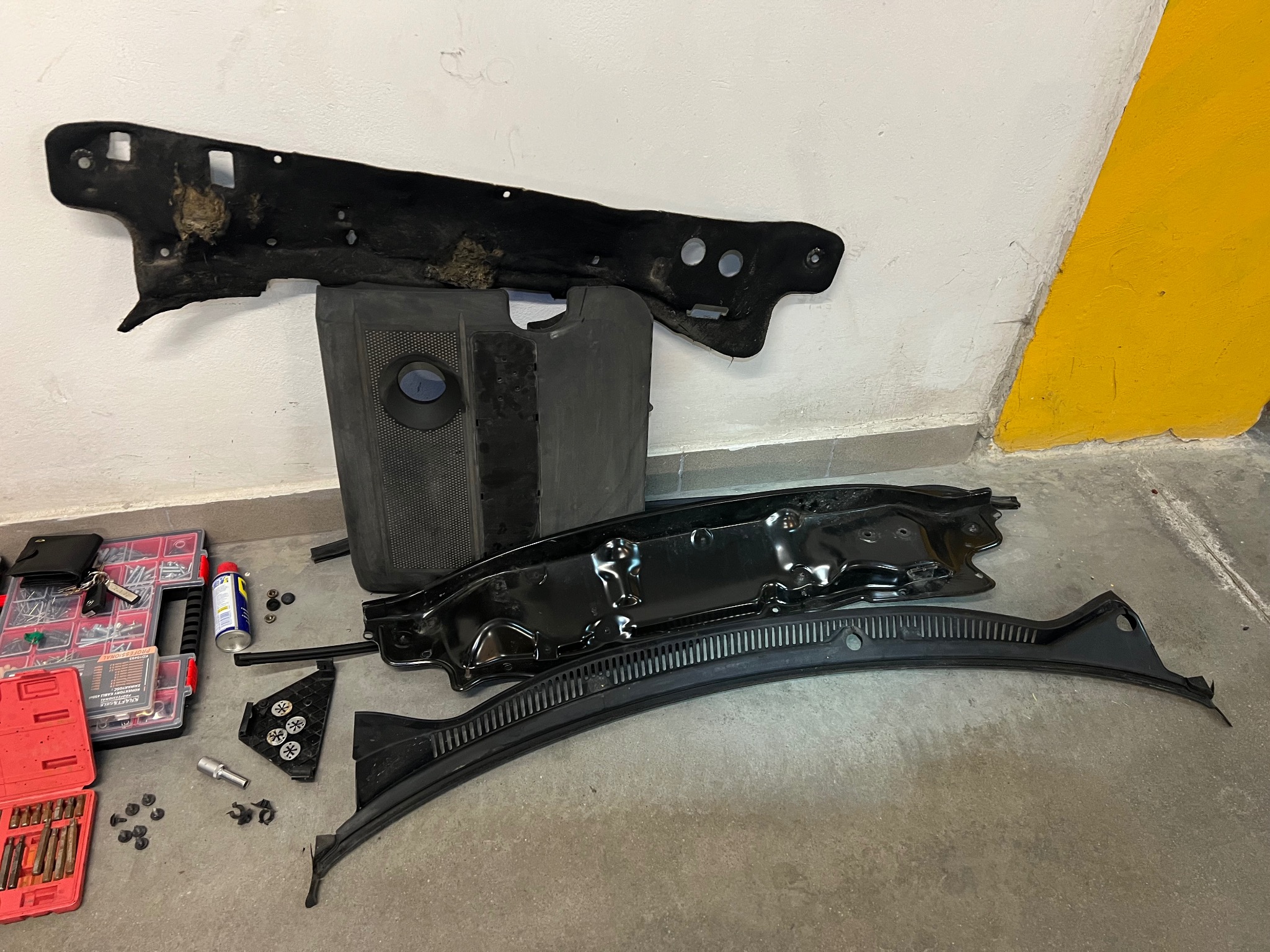 Demontaż elementów podszybia VW Polo: czarna listwa podszybia z widocznymi otworami, osłona filtra kabinowego, metalowa rama podszybia, skrzynka z narzędziami, klucze, śruby i spinki na betonowej...