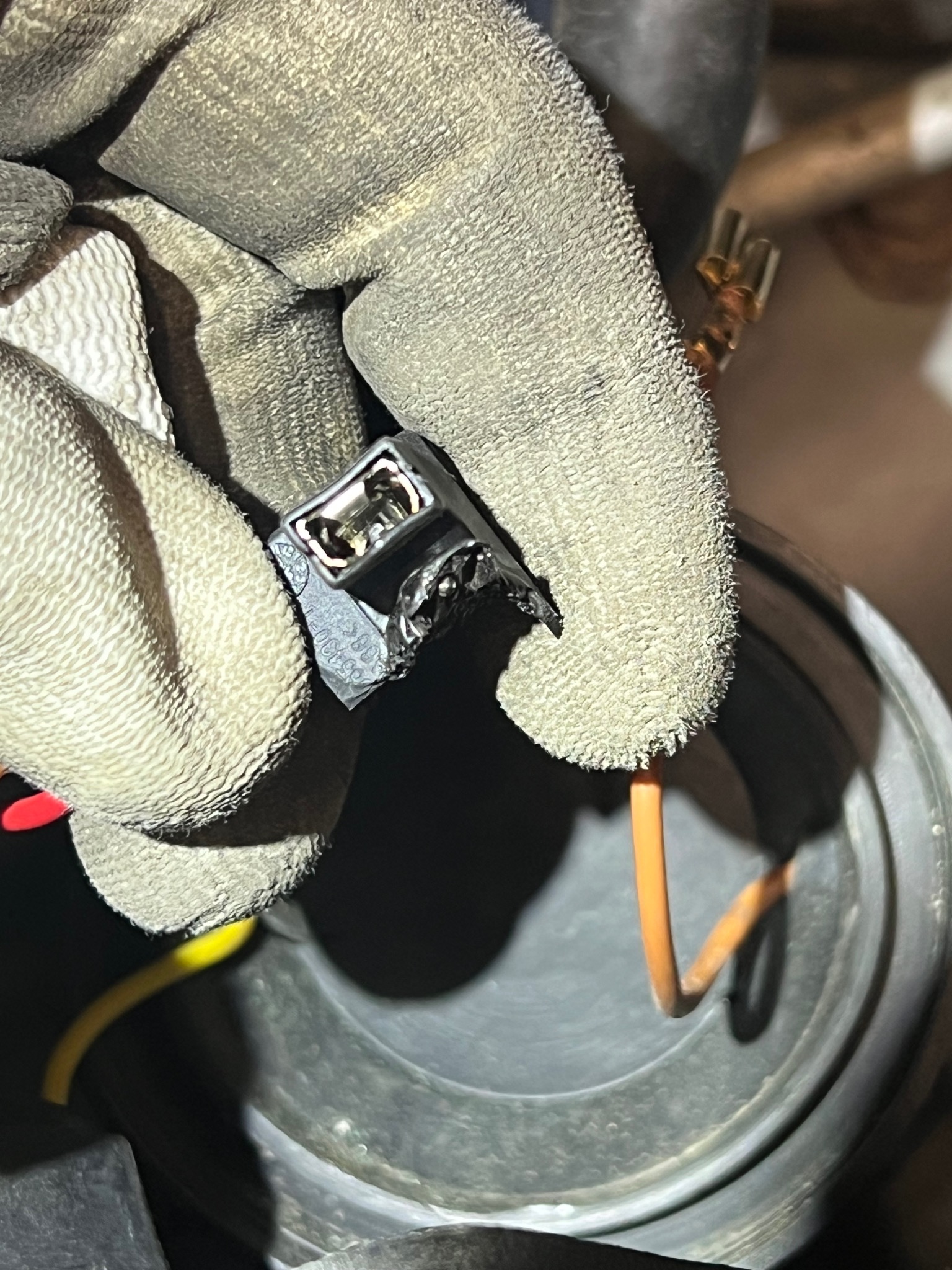 Dłoń w roboczej rękawicy trzyma czarną kostkę elektryczną z widocznymi stykami, obok widoczne przewody: pomarańczowy i żółty, oraz fragment reflektora samochodowego.