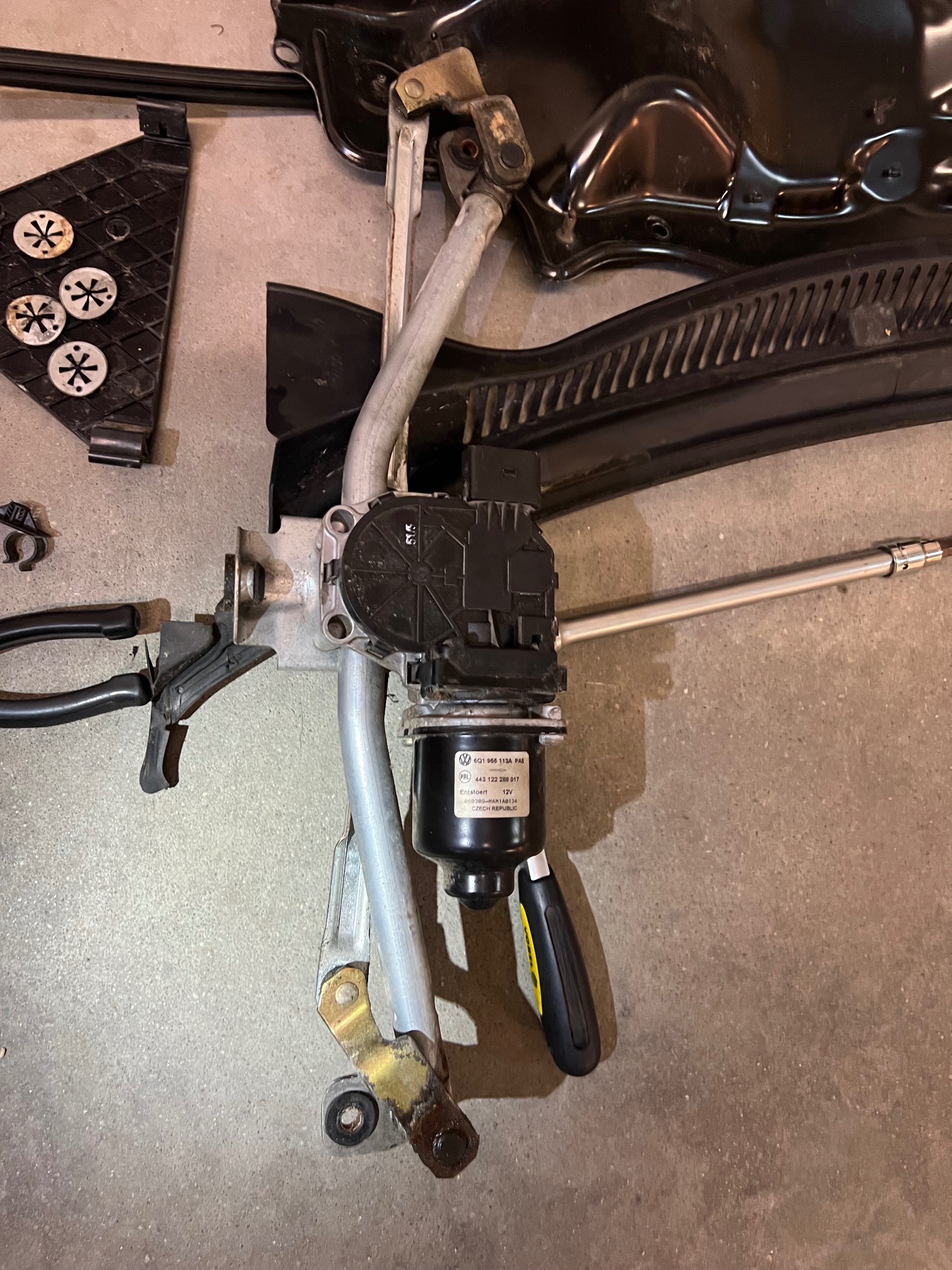 Zdemontowany mechanizm wycieraczek samochodowych VW Polo z widocznym silniczkiem, cięgnami, elementami mocującymi i narzędziem, leżący na szarej powierzchni.