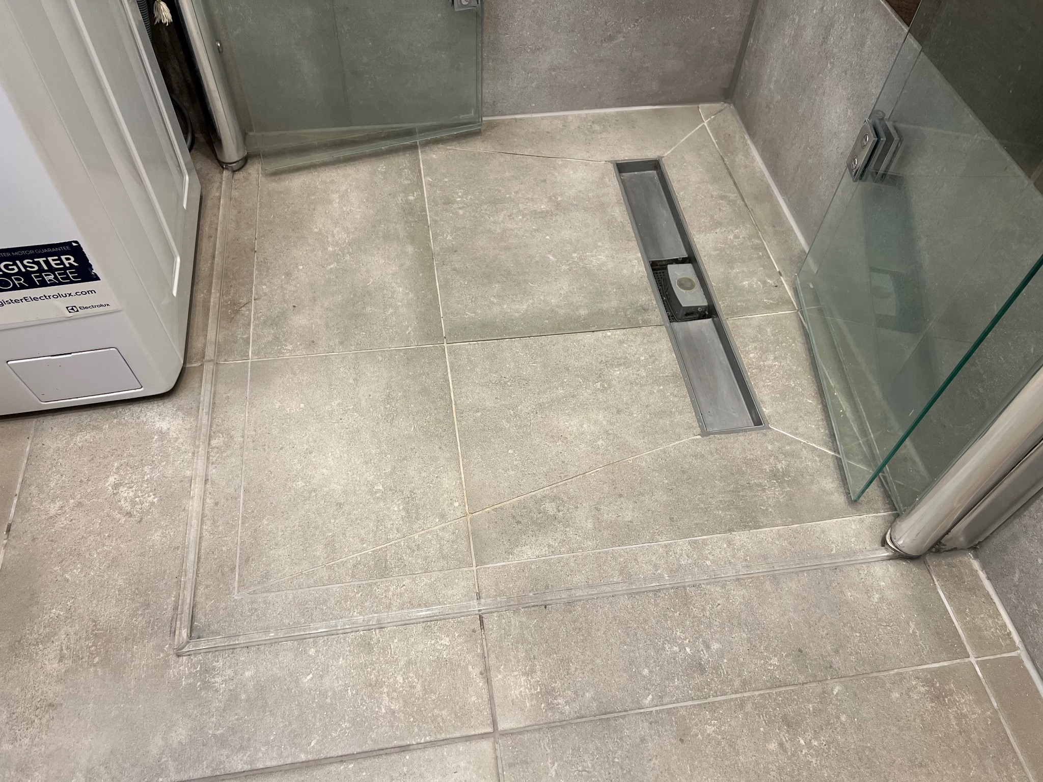 Zamontowany przezroczysty próg akrylowy brodzika z odpływem liniowym, uszczelniony szarym silikonem dopasowanym do fug, widoczne płytki podłogowe i fragment kabiny prysznicowej.