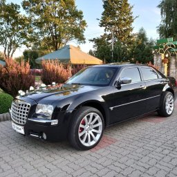 Przemysław Iwański - Czarny Chrysler 300C udekorowany białymi kwiatami na masce, zaparkowany na brukowanym placu przed restauracją z zielonymi parasolami.