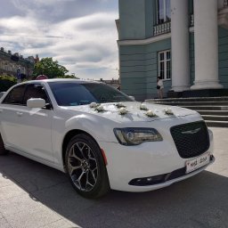 Przemysław Iwański - Biały Chrysler 300 udekorowany białymi różami na masce, z tablicą 'Mąż i Żona', zaparkowany przed budynkiem z kolumnami.