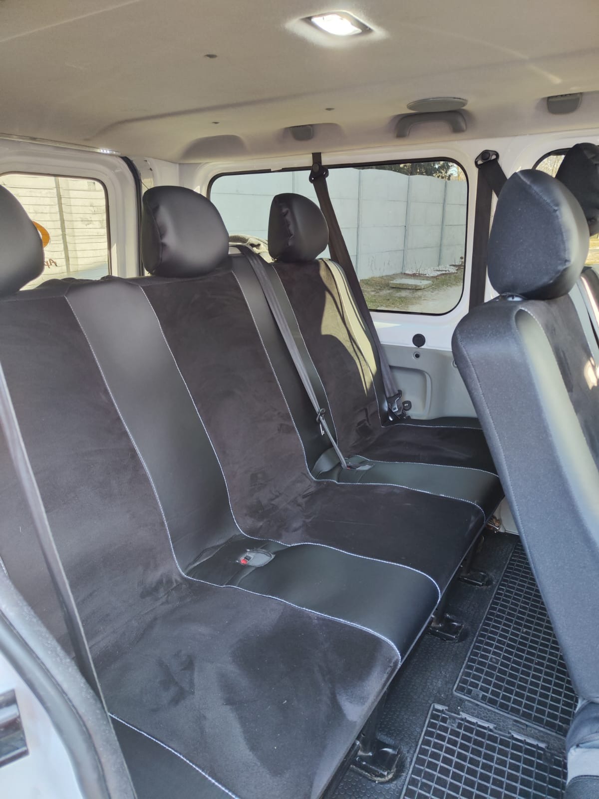 Wnętrze białego busa z trzema rzędami czarnych foteli z welurowymi wstawkami i białymi przeszyciami, widok z tyłu pojazdu.