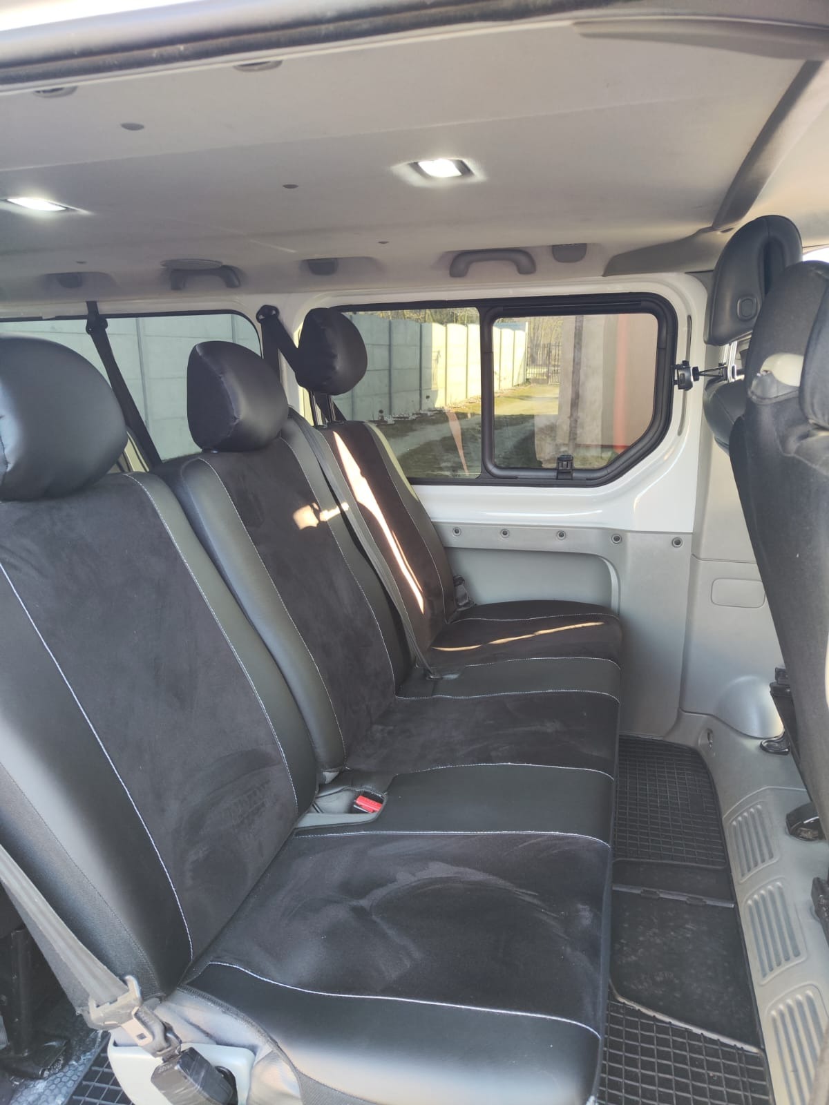 Wnętrze białego busa z trzema rzędami czarnych, welurowych siedzeń z białymi przeszyciami, widok z perspektywy pasażera, z widocznymi oknami i zagłówkami.