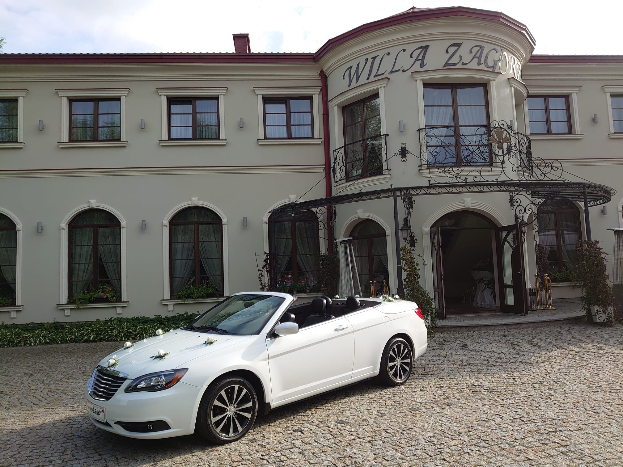Biały kabriolet udekorowany białymi kwiatami, zaparkowany przed budynkiem Willa Zagórze z ozdobnym wejściem.