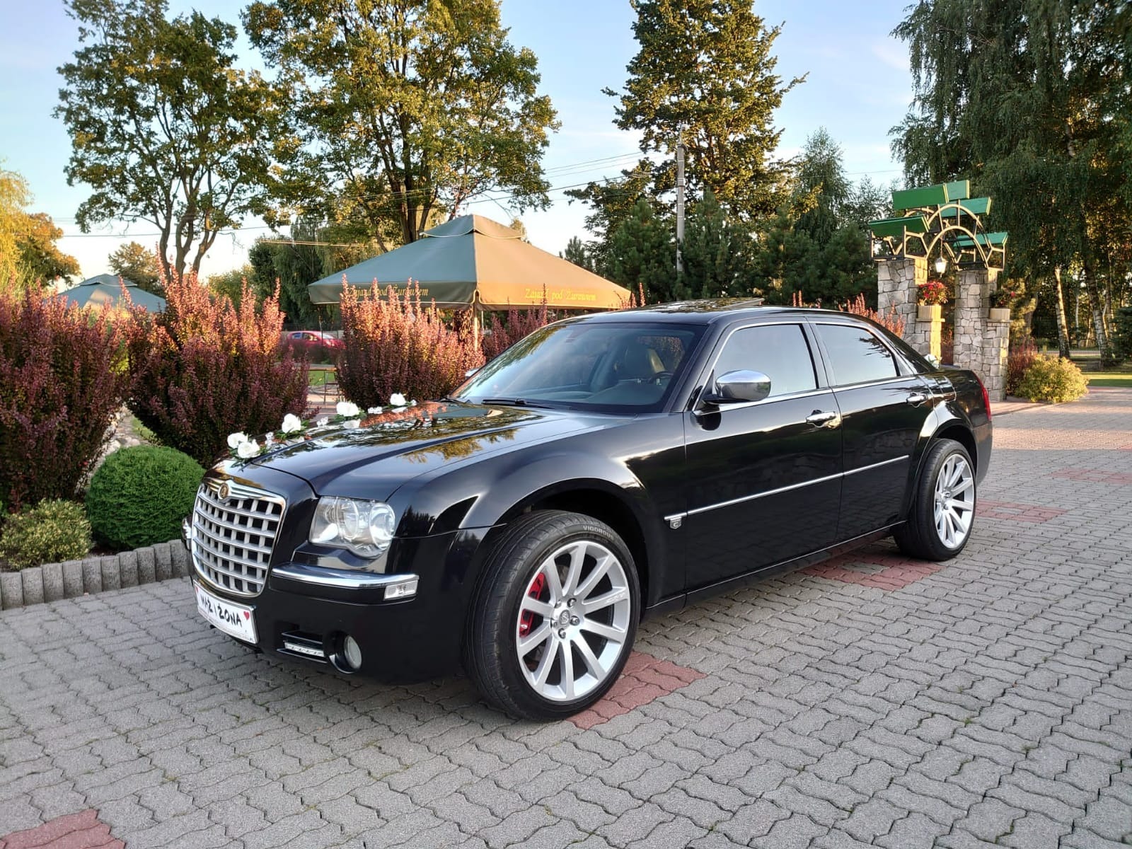 Czarny Chrysler 300C udekorowany białymi kwiatami na masce, zaparkowany na brukowanym placu przed restauracją z zielonymi parasolami.