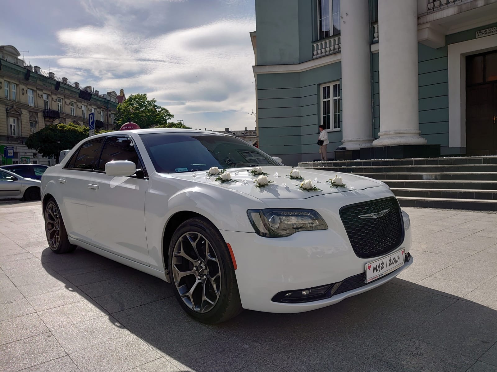 Biały Chrysler 300 udekorowany białymi różami na masce, z tablicą 'Mąż i Żona', zaparkowany przed budynkiem z kolumnami.
