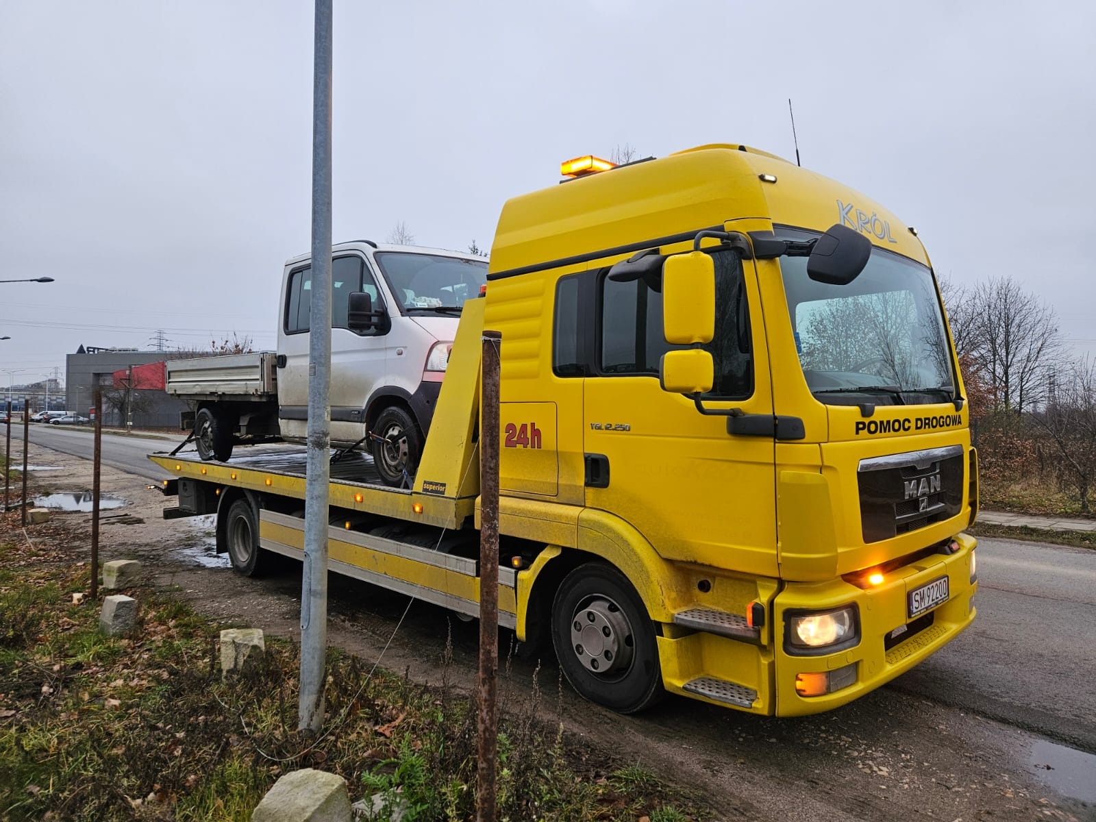 Żółty samochód pomocy drogowej marki MAN z napisem 'KRÓL' na dachu, transportujący biały samochód dostawczy na platformie, widok z boku na tle szarego nieba i zabudowań.