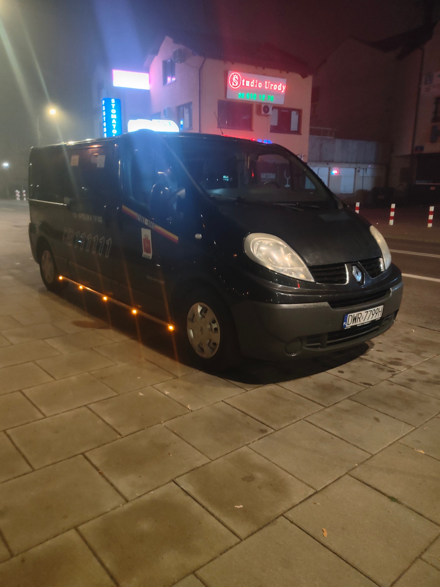 Czarny bus Renault Trafic z numerem telefonu i bocznymi światłami pozycyjnymi na tle nocnego krajobrazu miejskiego z oświetlonymi budynkami