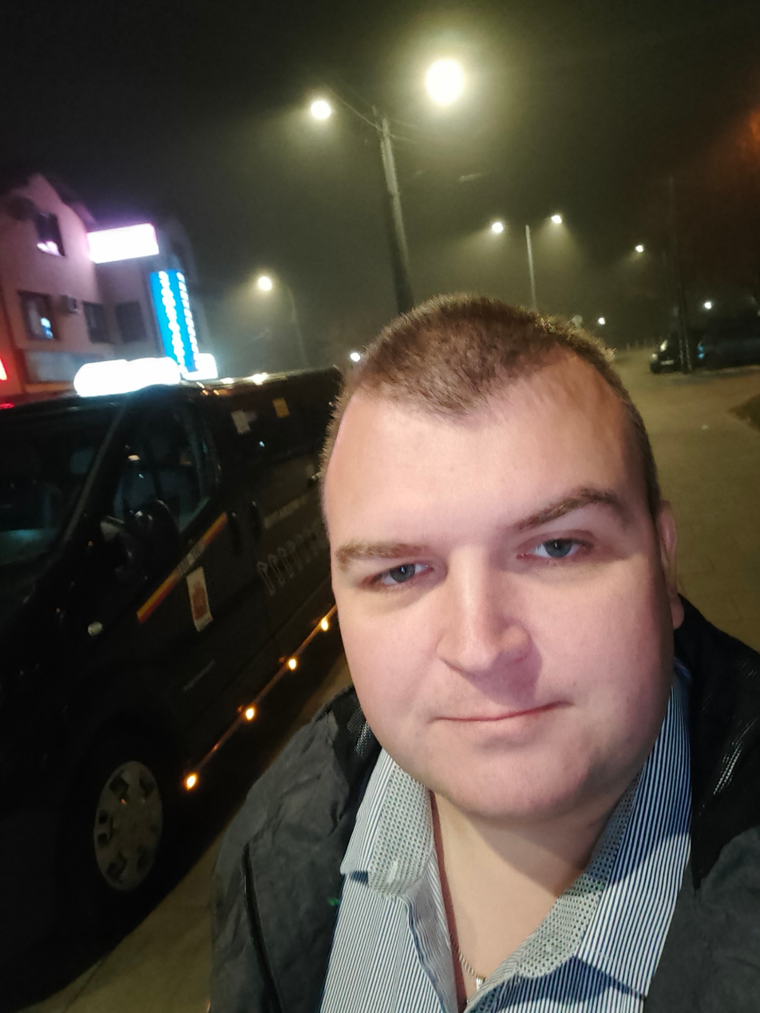 Mężczyzna robi selfie na tle czarnego busa z oświetleniem obrysowym i budynkiem w tle, nocna sceneria.
