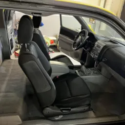 Wnętrze pickupa z widocznymi fotelami z czarną tapicerką, szarą wykładziną podłogową i deską rozdzielczą. Widoczny otwarty schowek i elementy wyposażenia. Wnętrze wydaje się być po praniu, ze...