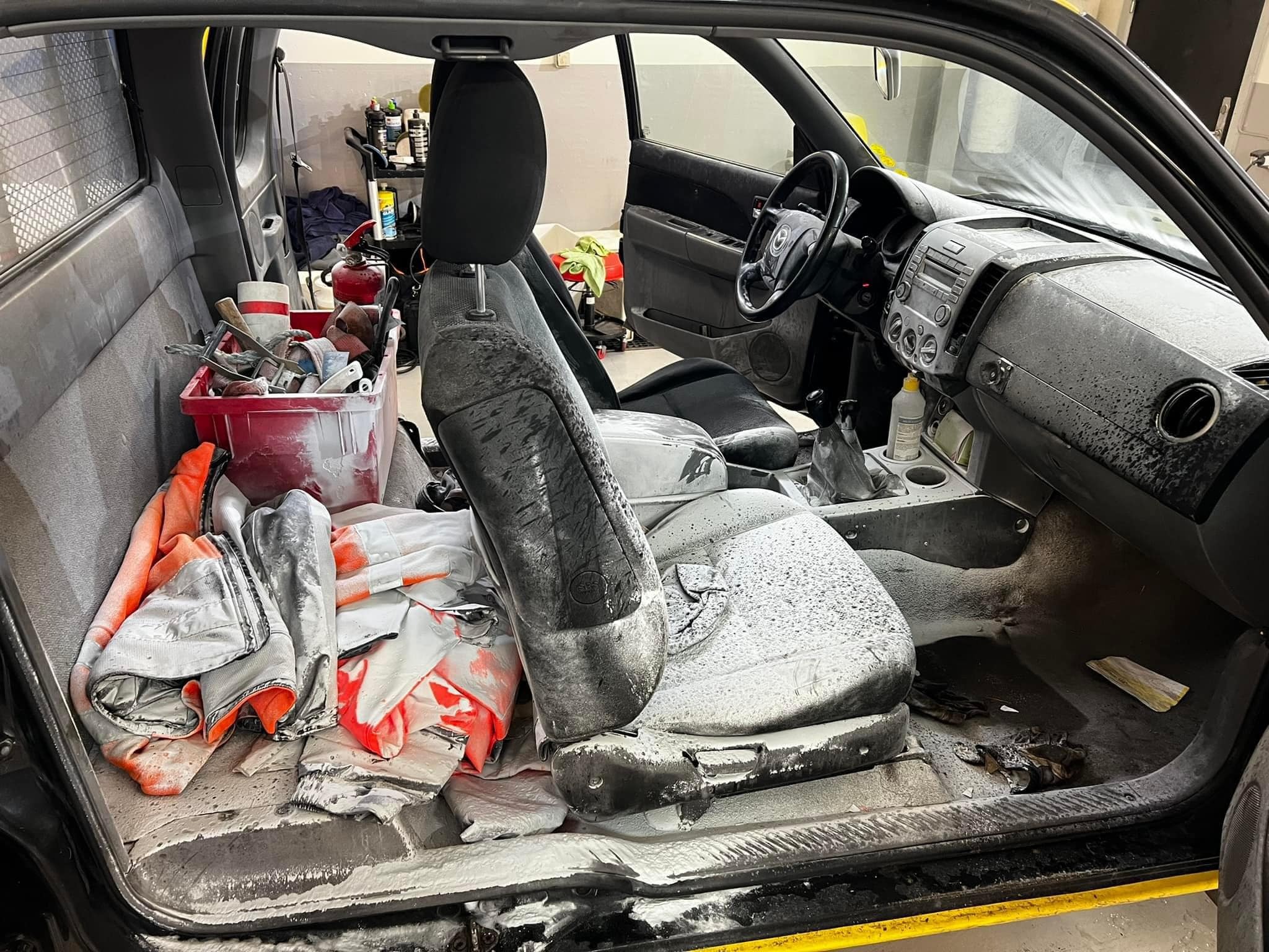 Wnętrze samochodu po czyszczeniu tapicerki z widocznymi pozostałościami środka czyszczącego na siedzeniach, desce rozdzielczej i podłodze, z otwartymi drzwiami i narzędziami w tle.
