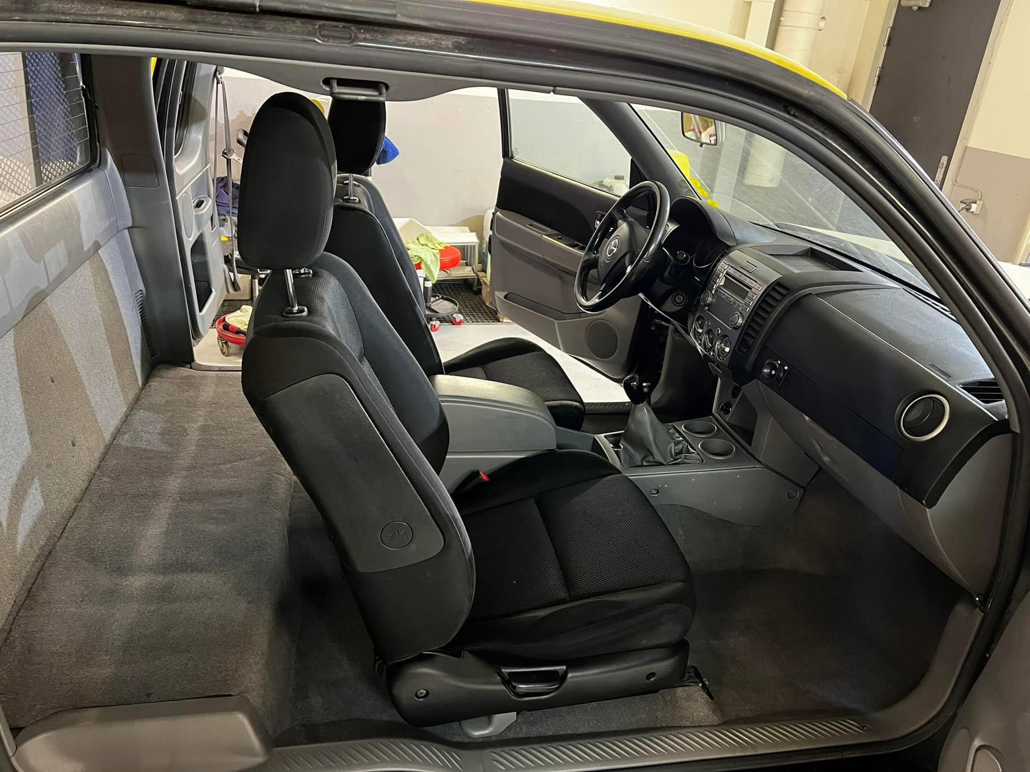 Wnętrze pickupa z widocznymi fotelami z czarną tapicerką, szarą wykładziną podłogową i deską rozdzielczą. Widoczny otwarty schowek i elementy wyposażenia. Wnętrze wydaje się być po praniu, ze...