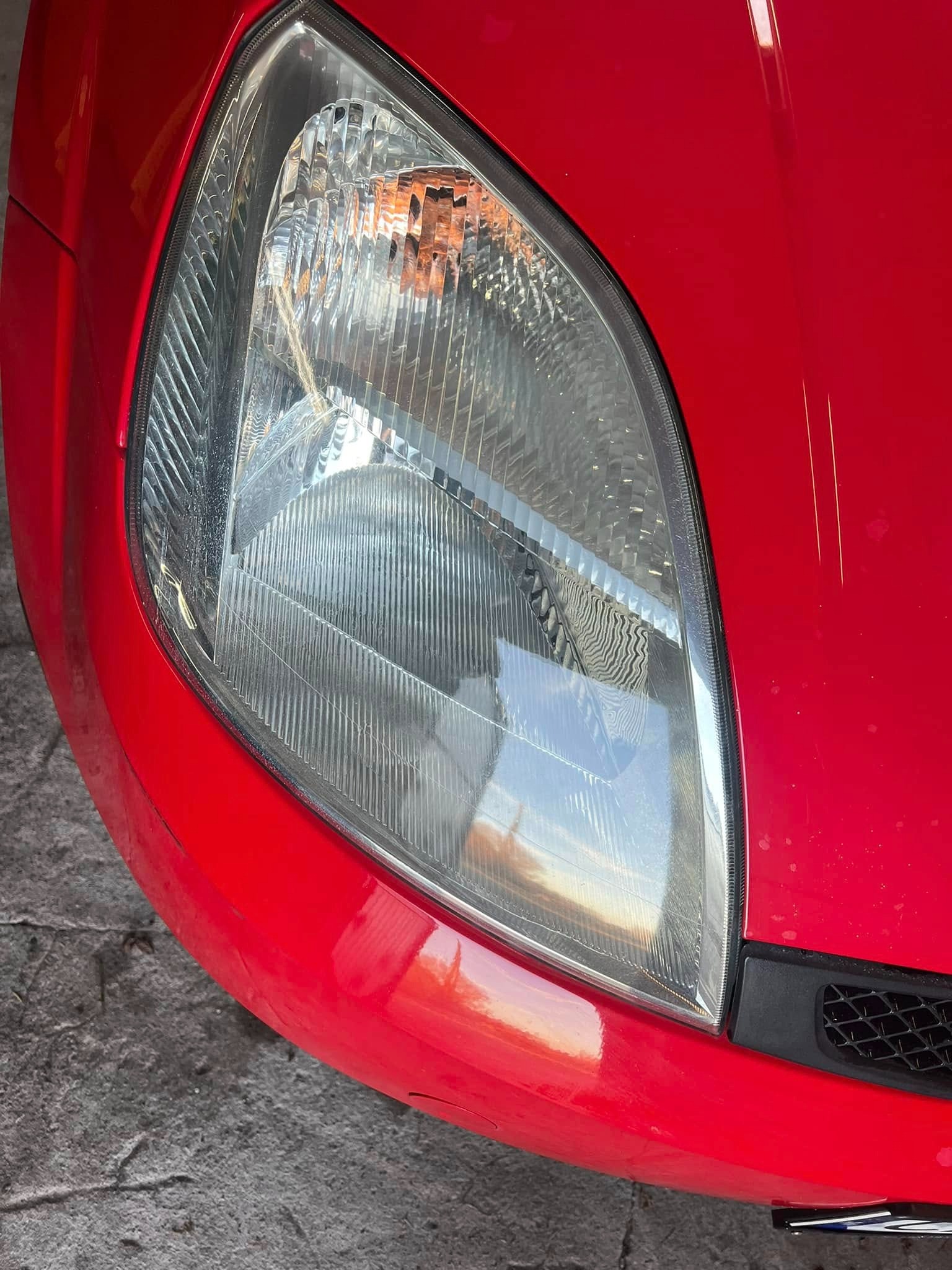 Zbliżenie na przedni reflektor i fragment czerwonego błotnika samochodu, widoczne drobne rysy na lakierze.