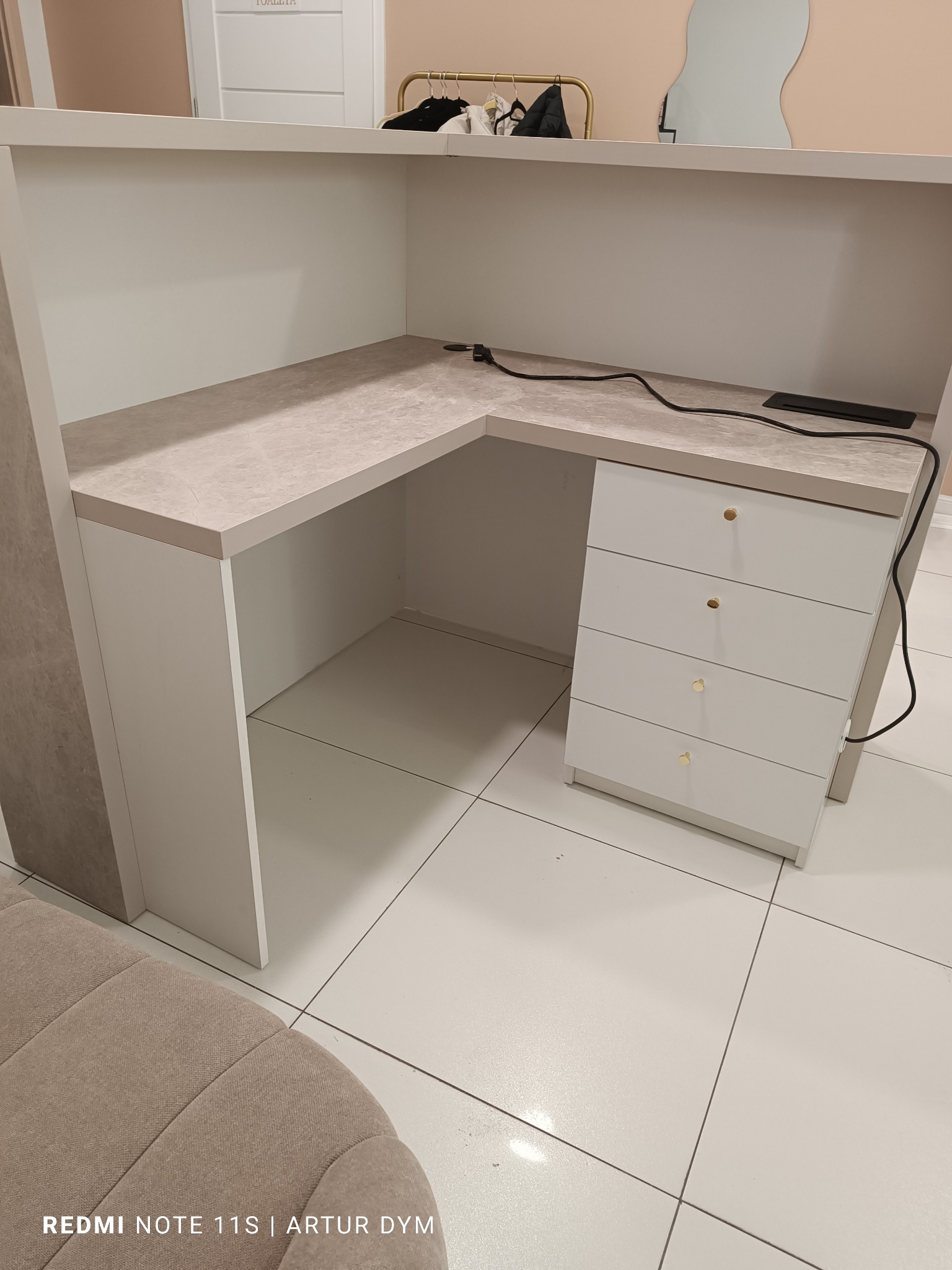 Narożne biurko z szufladami, wykonane na wymiar w Kielcach, z blatem imitującym marmur i złotymi gałkami. Widoczny kabel zasilający i fragment sofy.