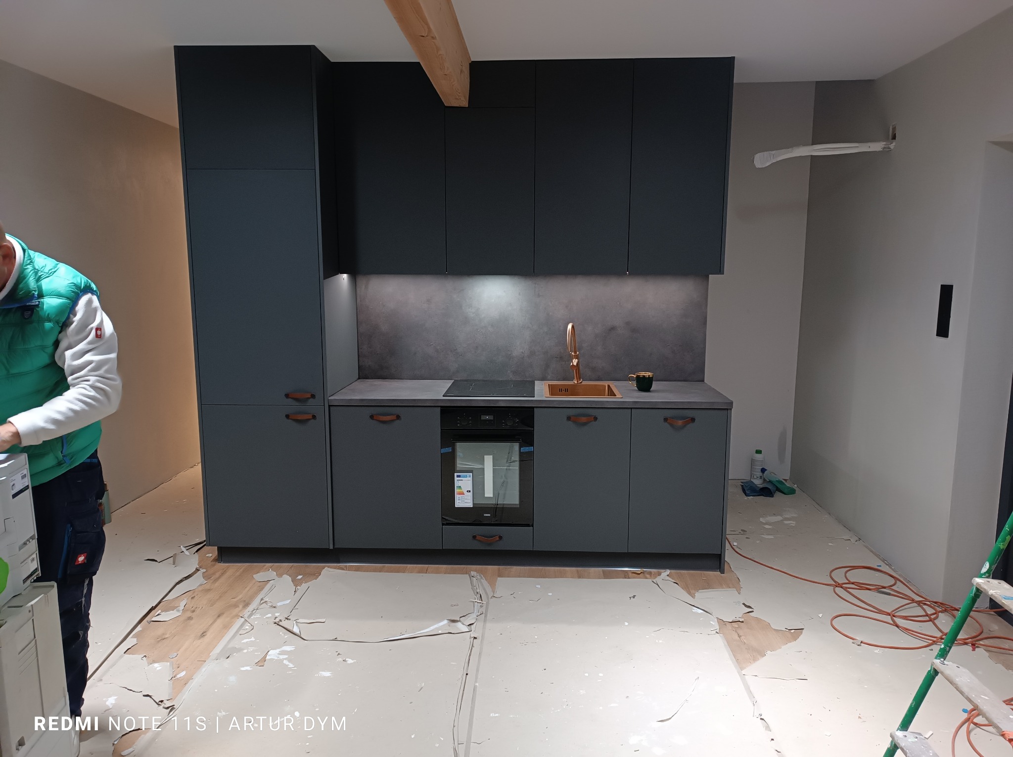Ciemnoszara zabudowa kuchenna z miedzianą armaturą i drewnianymi uchwytami, widoczna w trakcie prac wykończeniowych, z częściowo zabezpieczoną podłogą i fragmentem drabiny malarskiej.