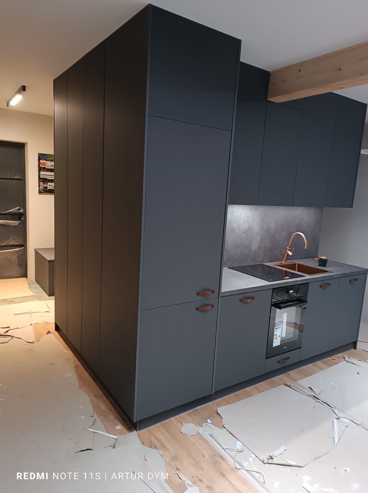 Ciemnoszara zabudowa kuchenna z szafkami podwieszanymi, piekarnikiem i złotą baterią, widoczne ślady montażu na podłodze.