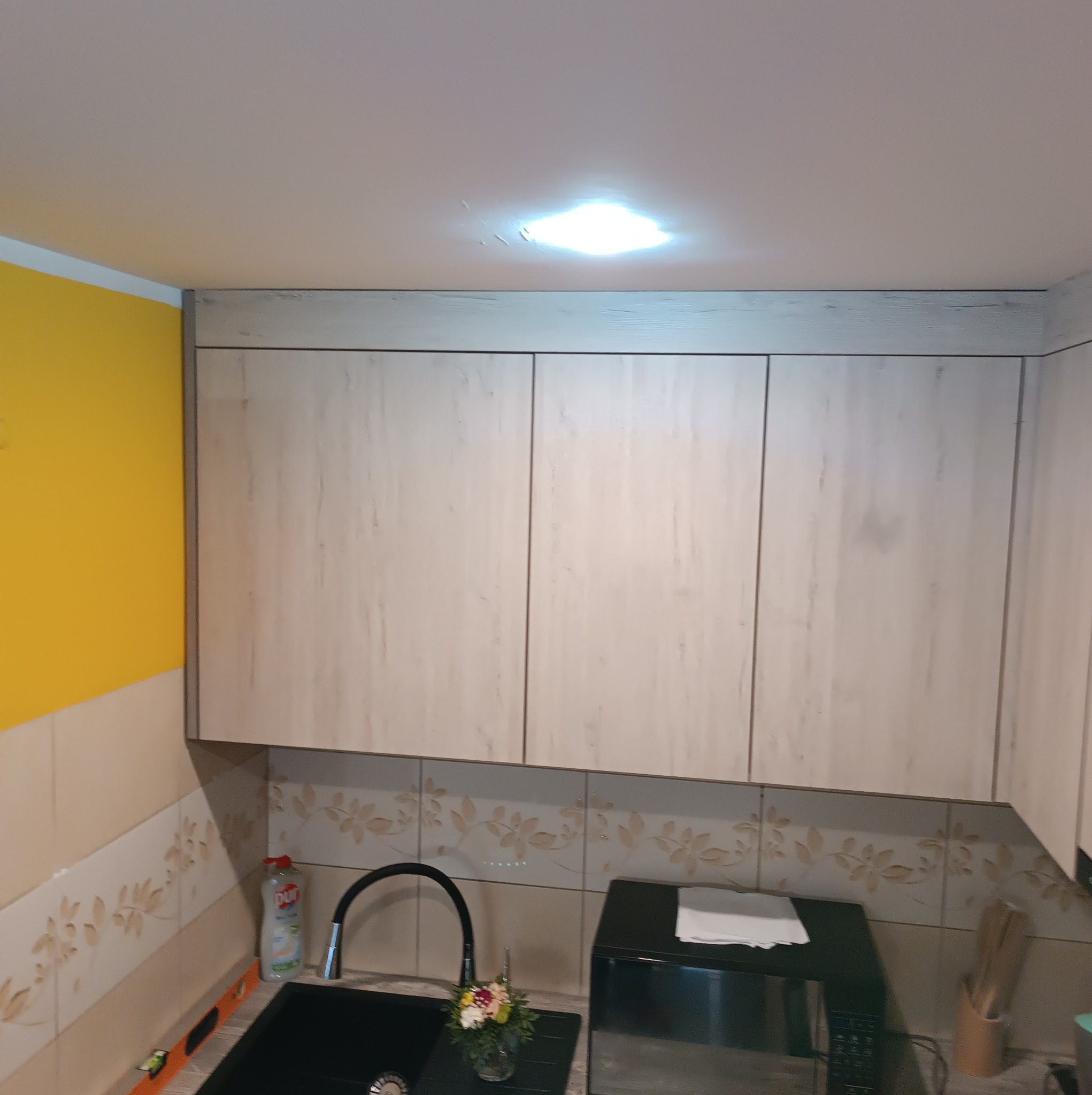 Zamontowane szafki kuchenne w kolorze jasnego drewna, nad blatem z czarnym zlewem i małym bukietem kwiatów, żółta ściana po lewej stronie.