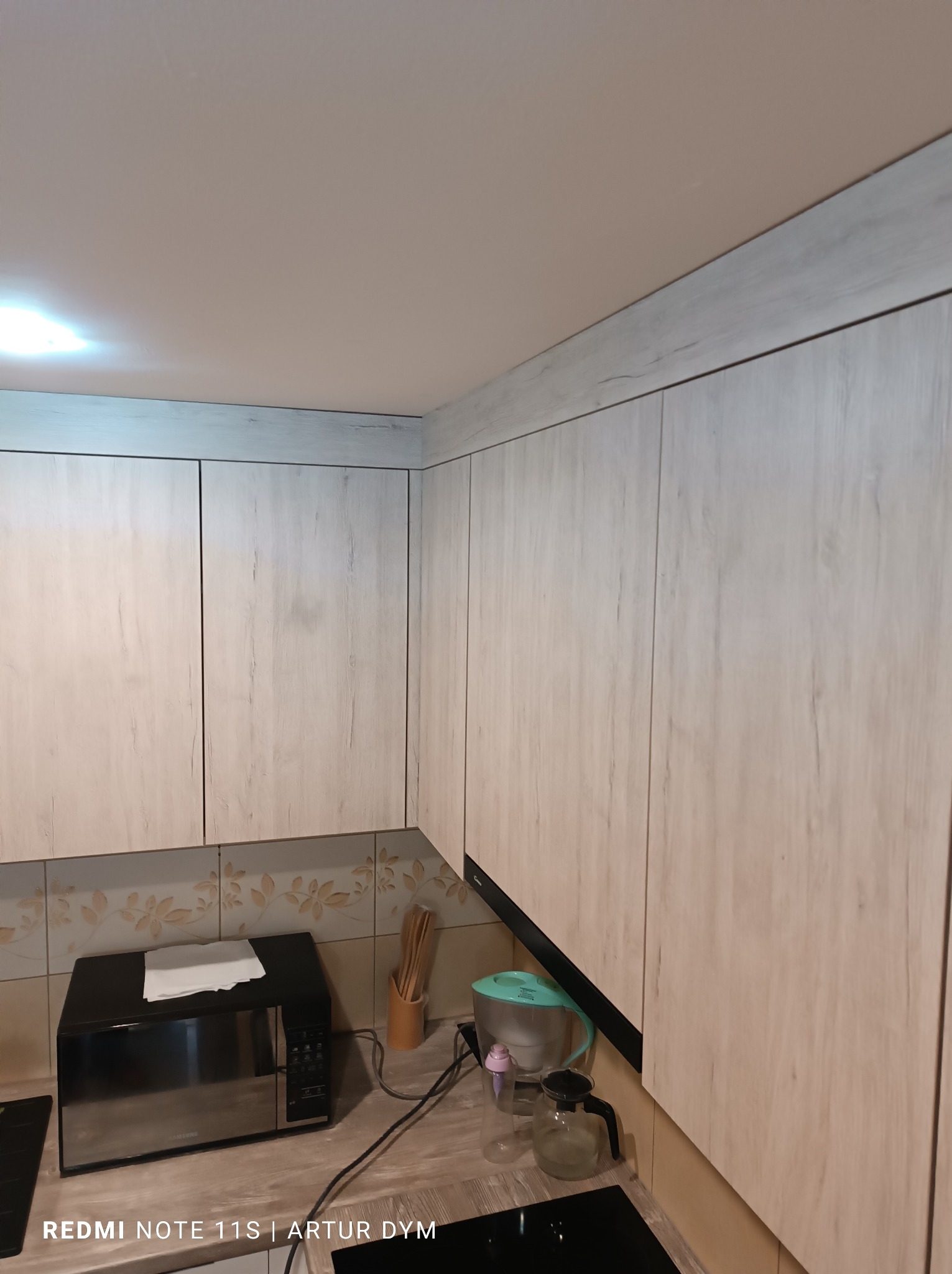 Jasne, drewnopodobne szafki kuchenne z mikrofala i akcesoriami na blacie, widok z dołu, oświetlenie sufitowe.