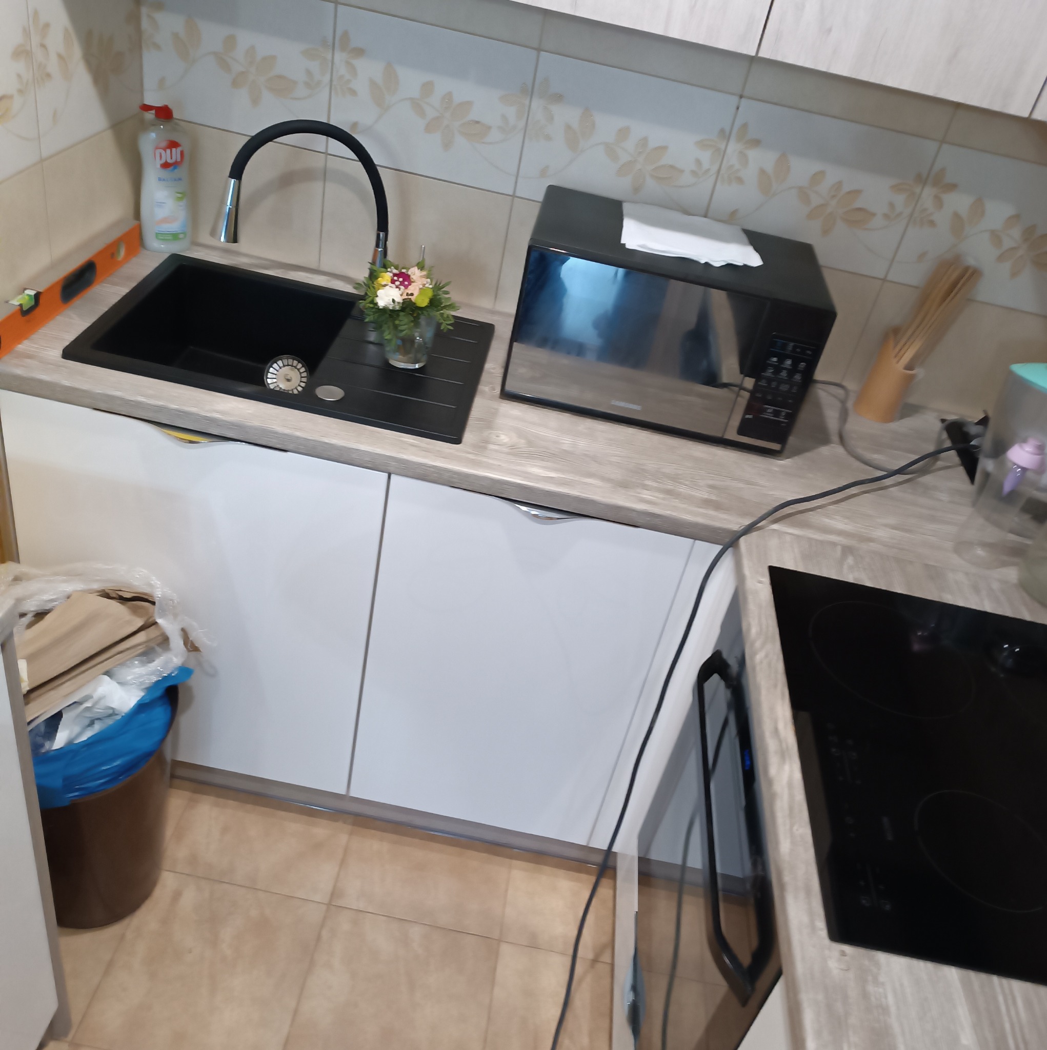 Nowoczesna kuchnia z czarnym zlewem, baterią w kształcie łuku i kuchenką indukcyjną, uzupełniona mikrofalówką i kwiatami w wazonie.