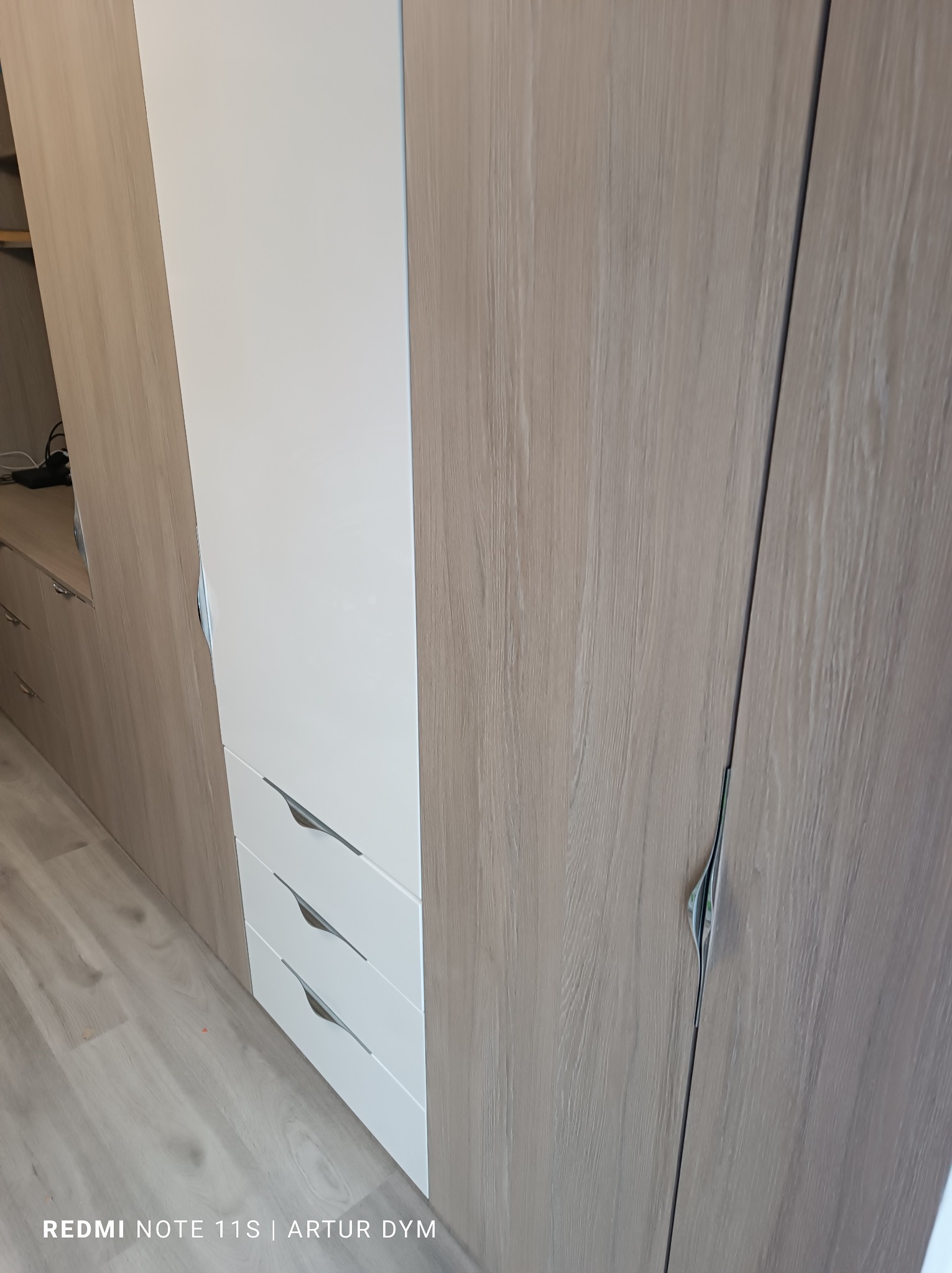 Fragment zabudowy meblowej z białymi szufladami i frontami w kolorze jasnego drewna, widoczne metalowe uchwyty szuflad.