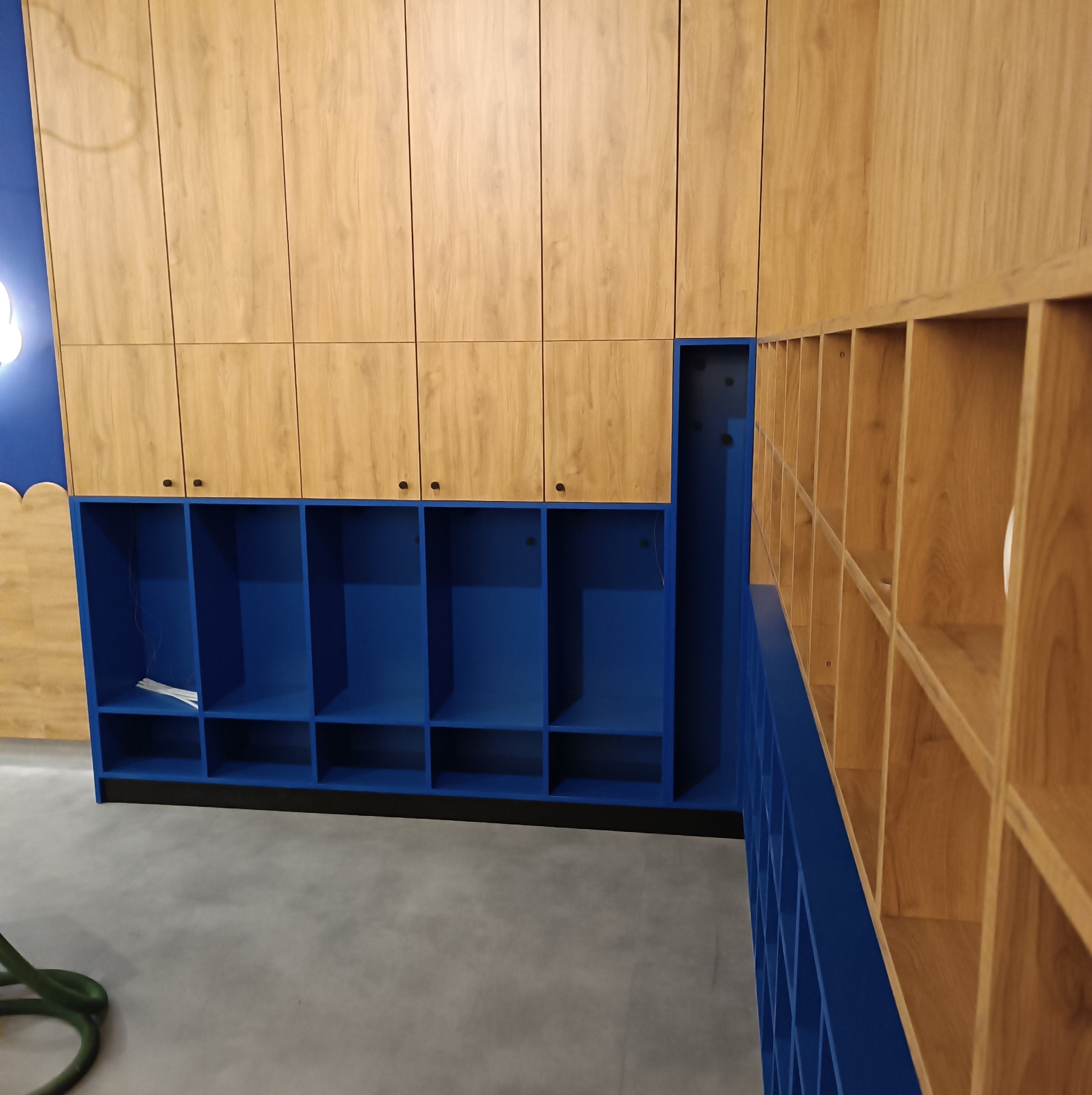 Niebiesko-drewniana zabudowa meblowa z szafkami i otwartymi półkami, widoczny fragment odkurzacza na podłodze.