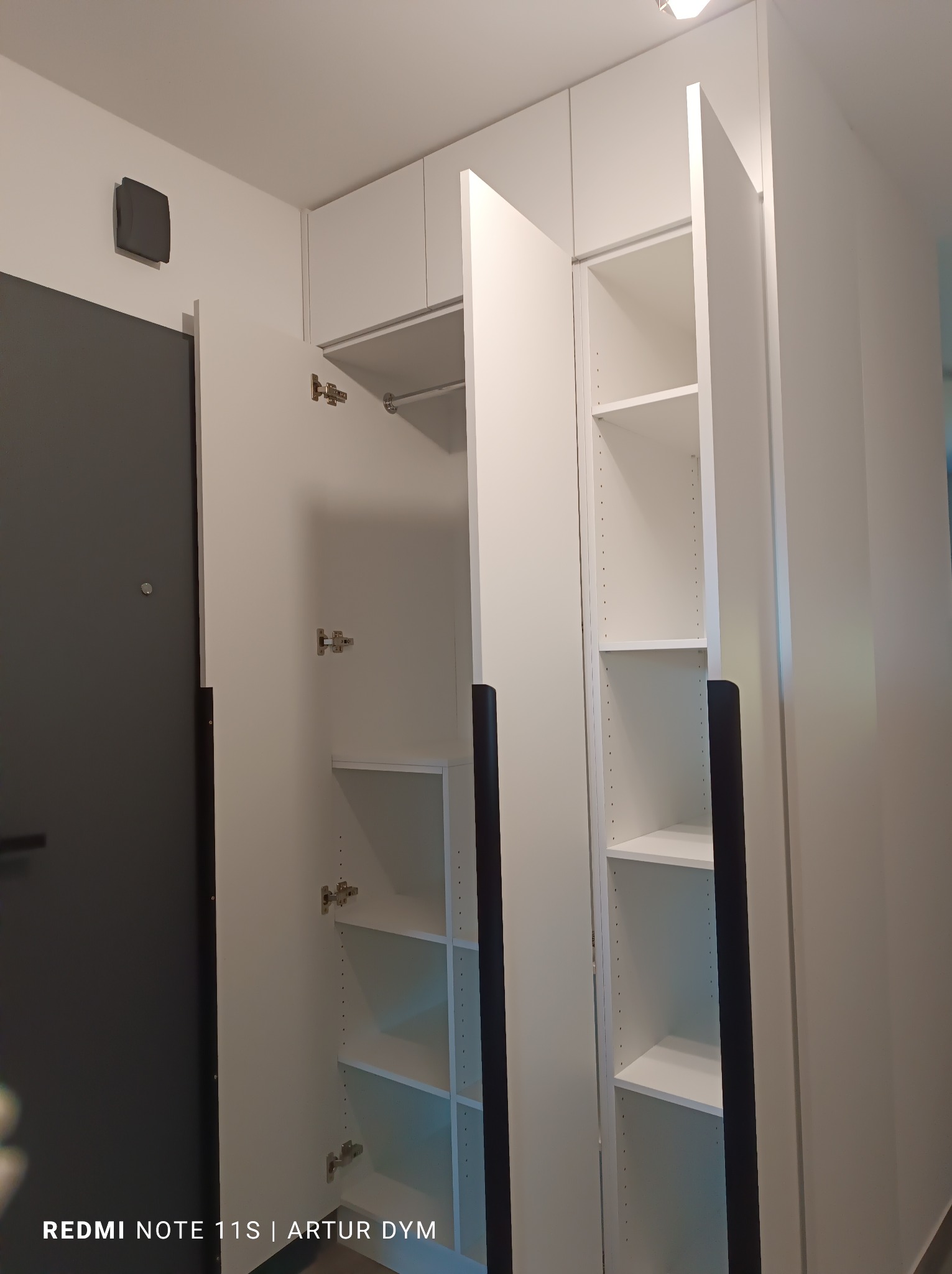 Biała szafa wnękowa z otwartymi drzwiami, widoczne półki i drążek na ubrania, czarne uchwyty, obok szare drzwi wejściowe.