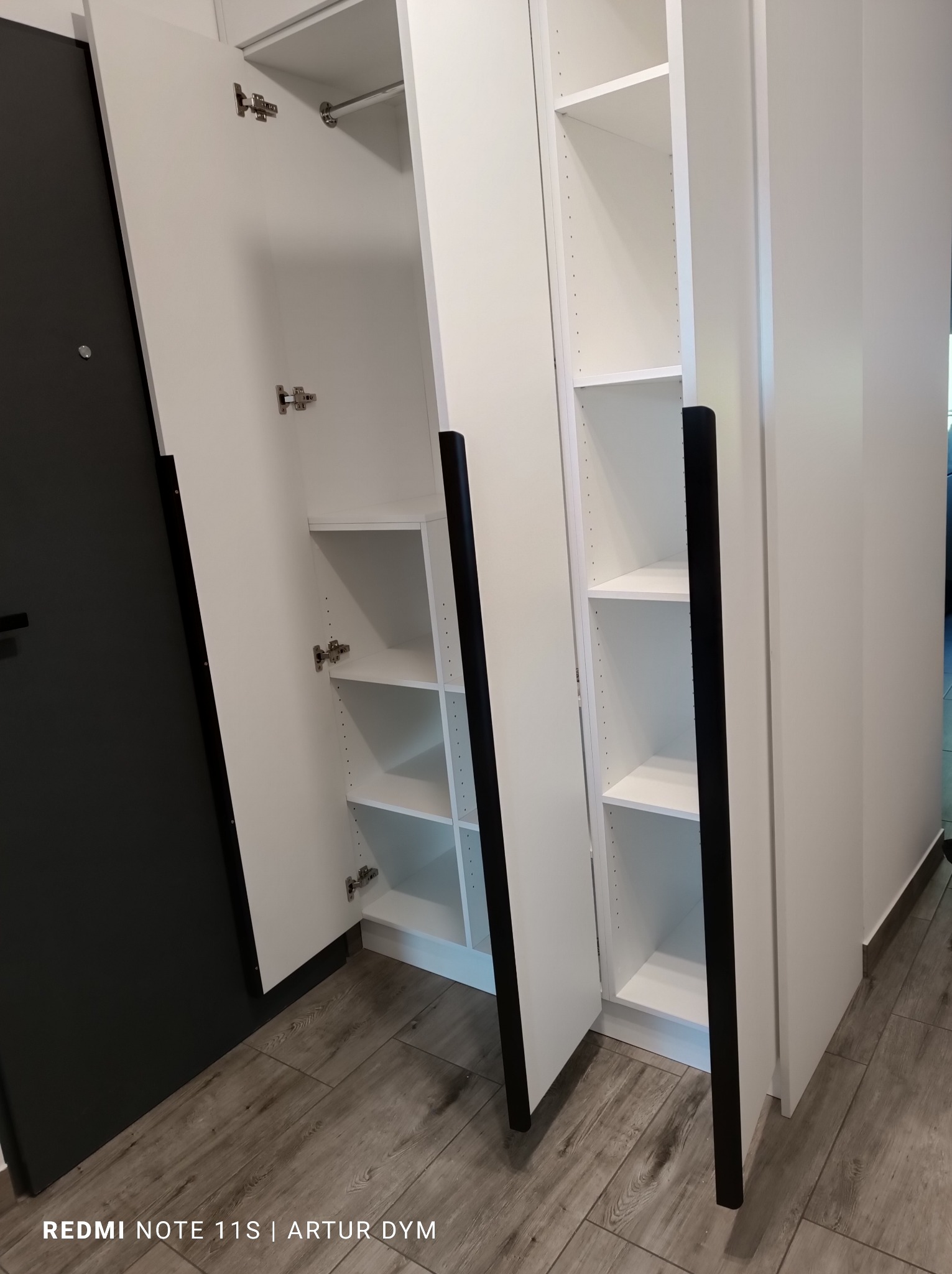 Biała szafa wnękowa z czarnymi, podłużnymi uchwytami, z otwartymi drzwiami ukazującymi puste półki, zamontowana przy ciemnych drzwiach wejściowych i drewnopodobnej podłodze.