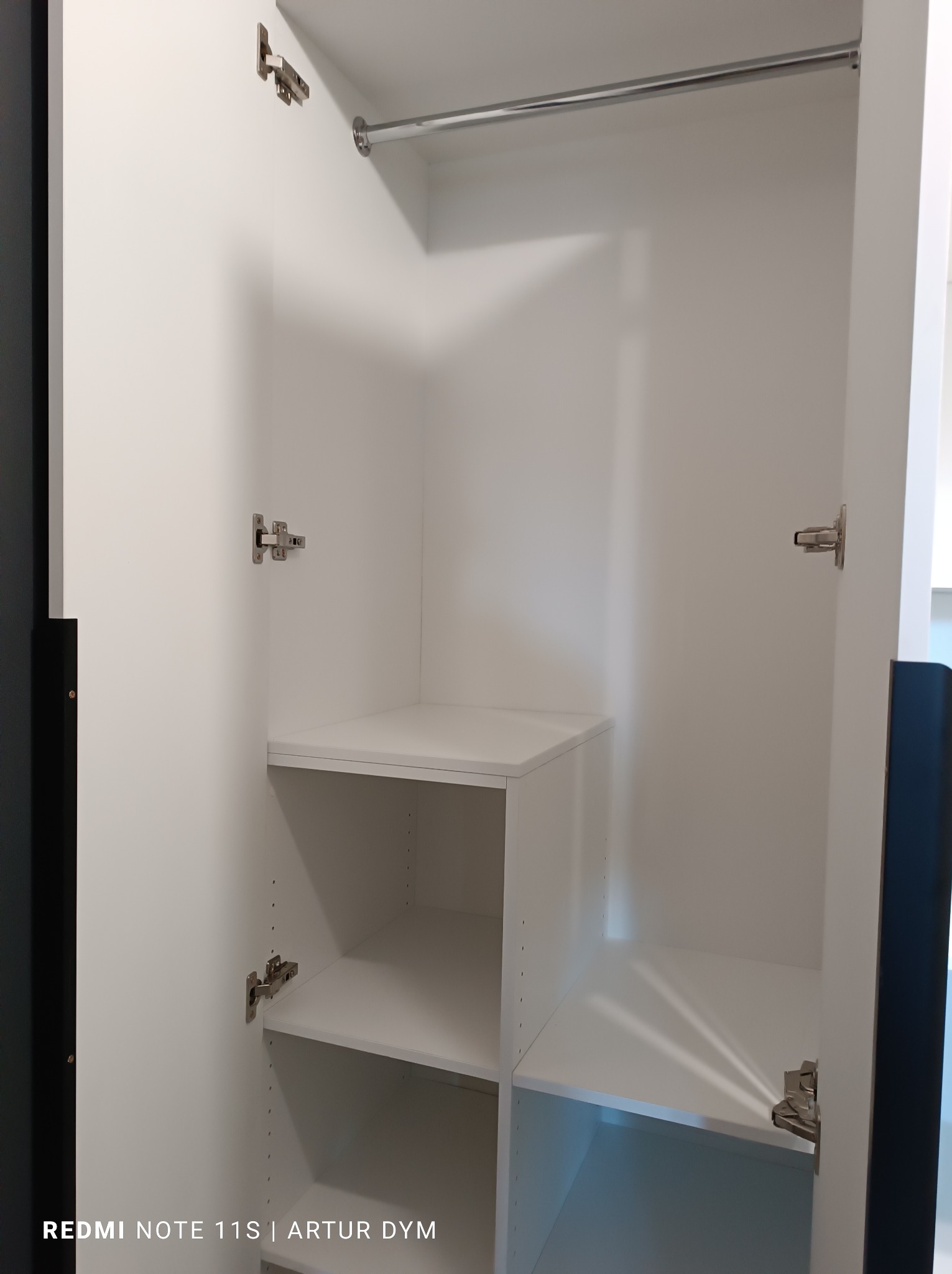 Wnętrze białej szafy z półkami i drążkiem, widoczne zawiasy i otwory na regulację półek.