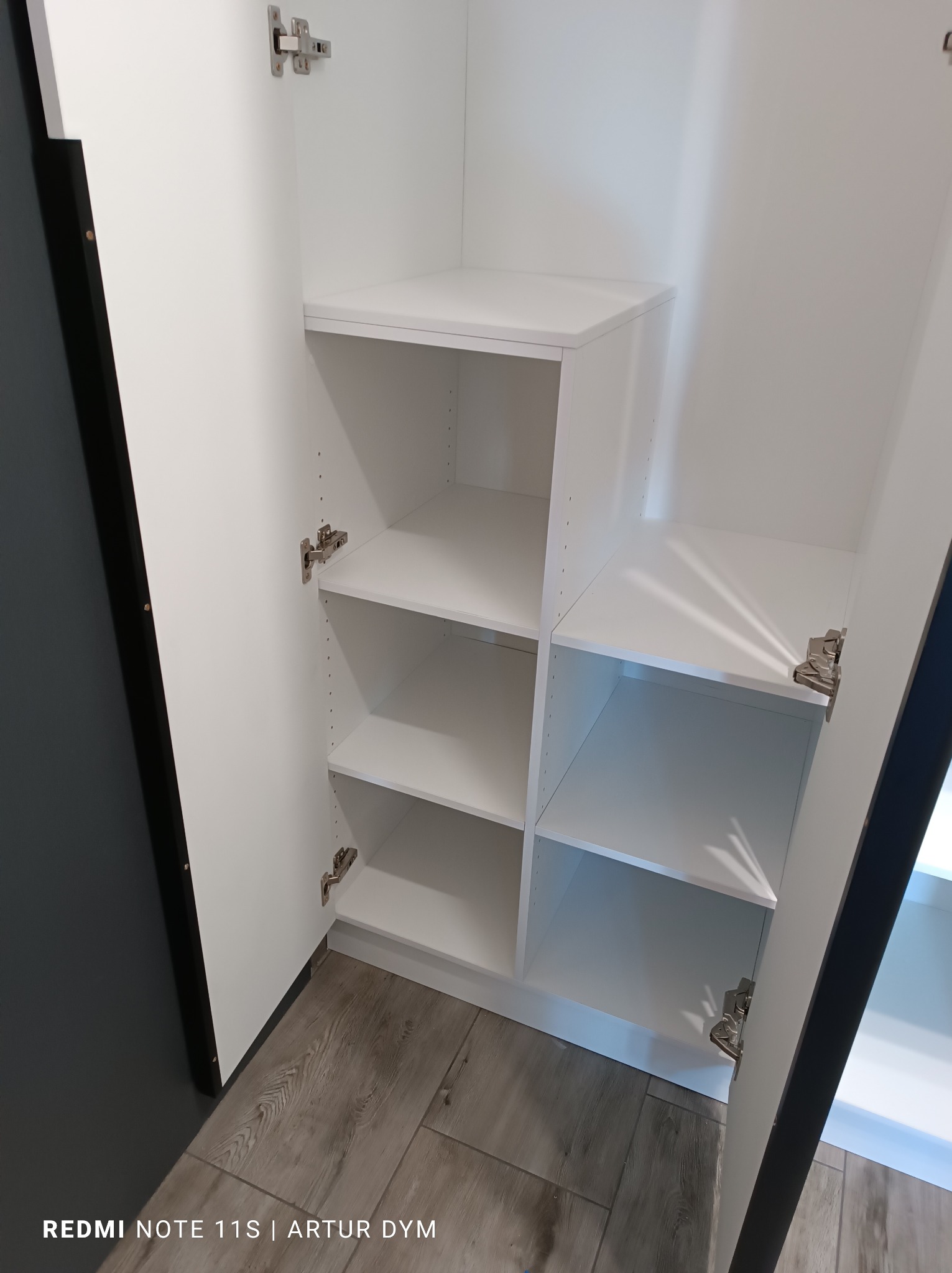 Wnętrze białej szafy z półkami, widok z otwartymi drzwiami, widoczne zawiasy i szara podłoga.