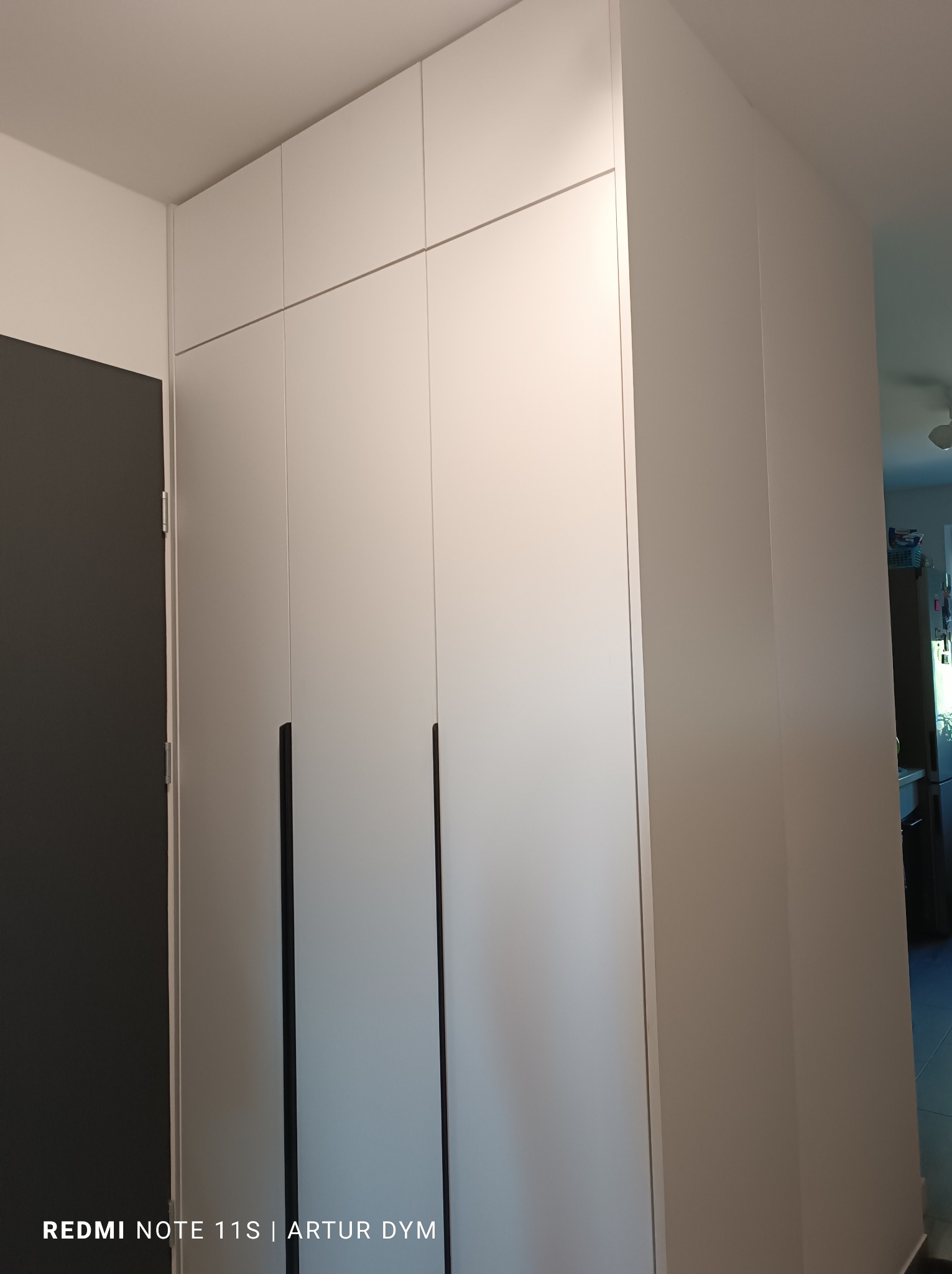 Biała szafa wnękowa z czarnymi, pionowymi uchwytami, widok z boku, z fragmentem szarej ściany po lewej stronie.