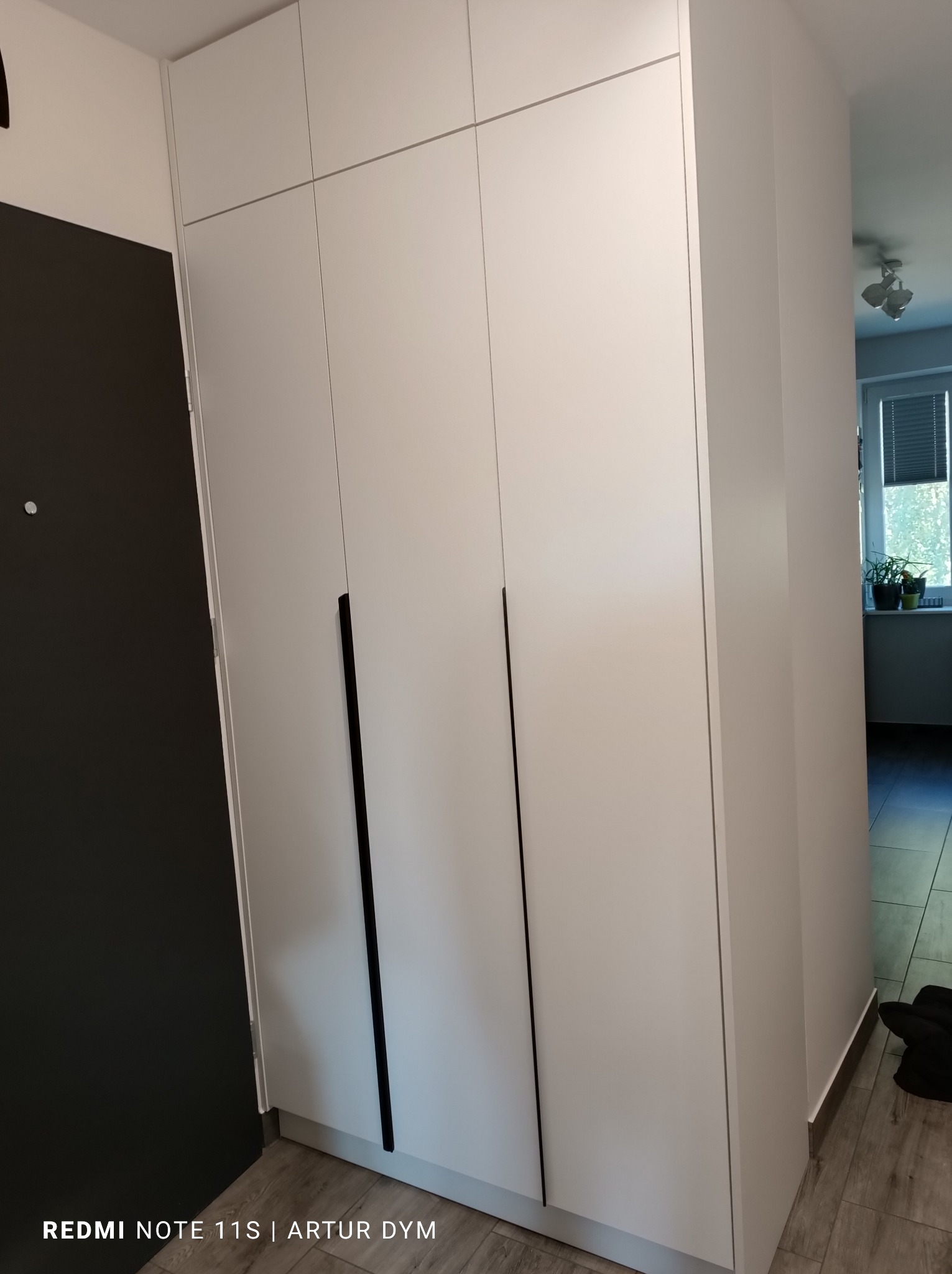 Wysoka, biała szafa wnękowa z czarnymi, minimalistycznymi uchwytami, umieszczona w przedpokoju przy czarnych drzwiach wejściowych.