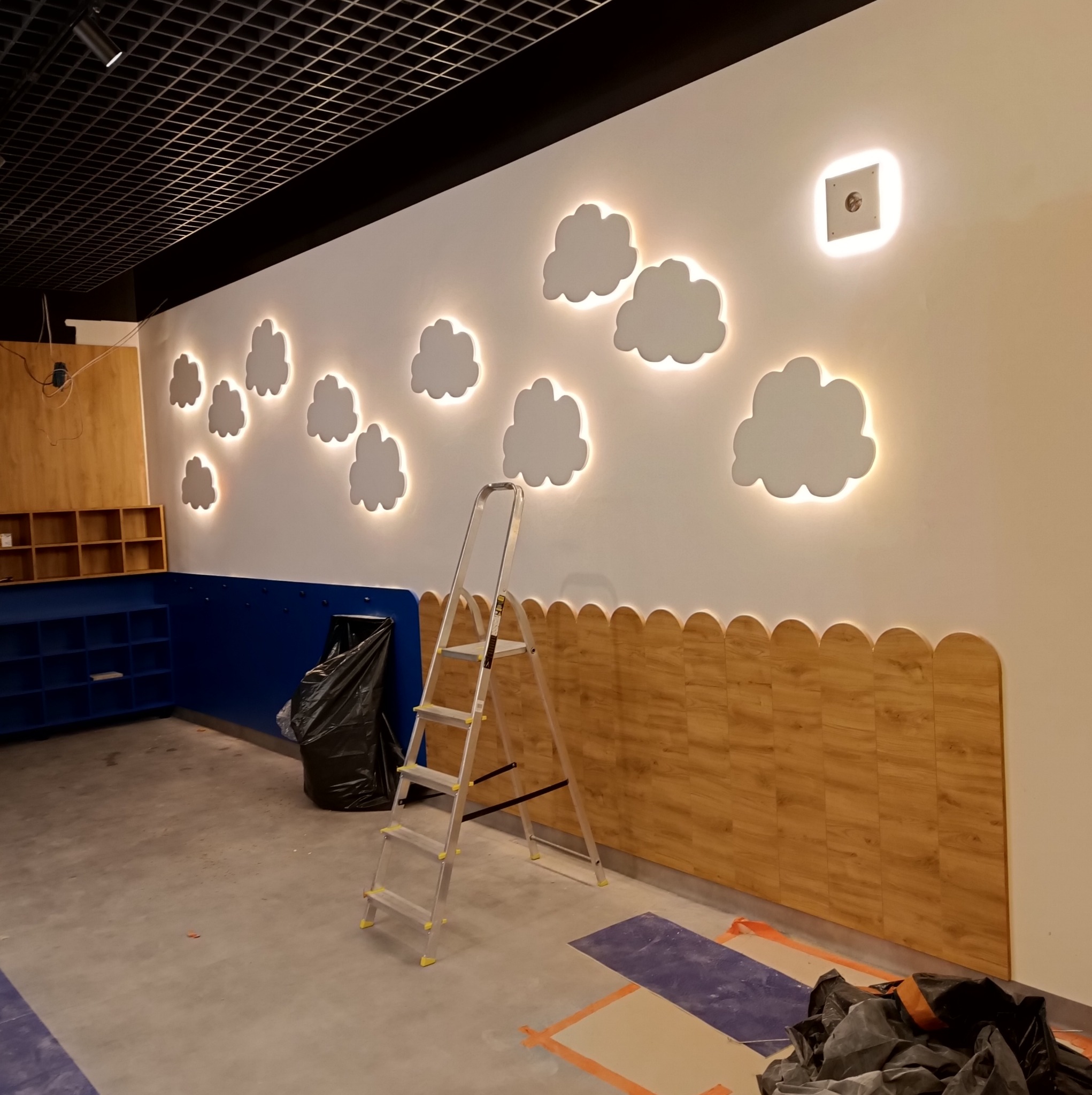 Pokój dziecięcy w trakcie wykańczania: zamontowane drewniane panele z zaokrąglonymi krawędziami na dole ściany, dekoracyjne chmurki z podświetleniem LED na górze, drabina oparta o ścianę, widoczny...