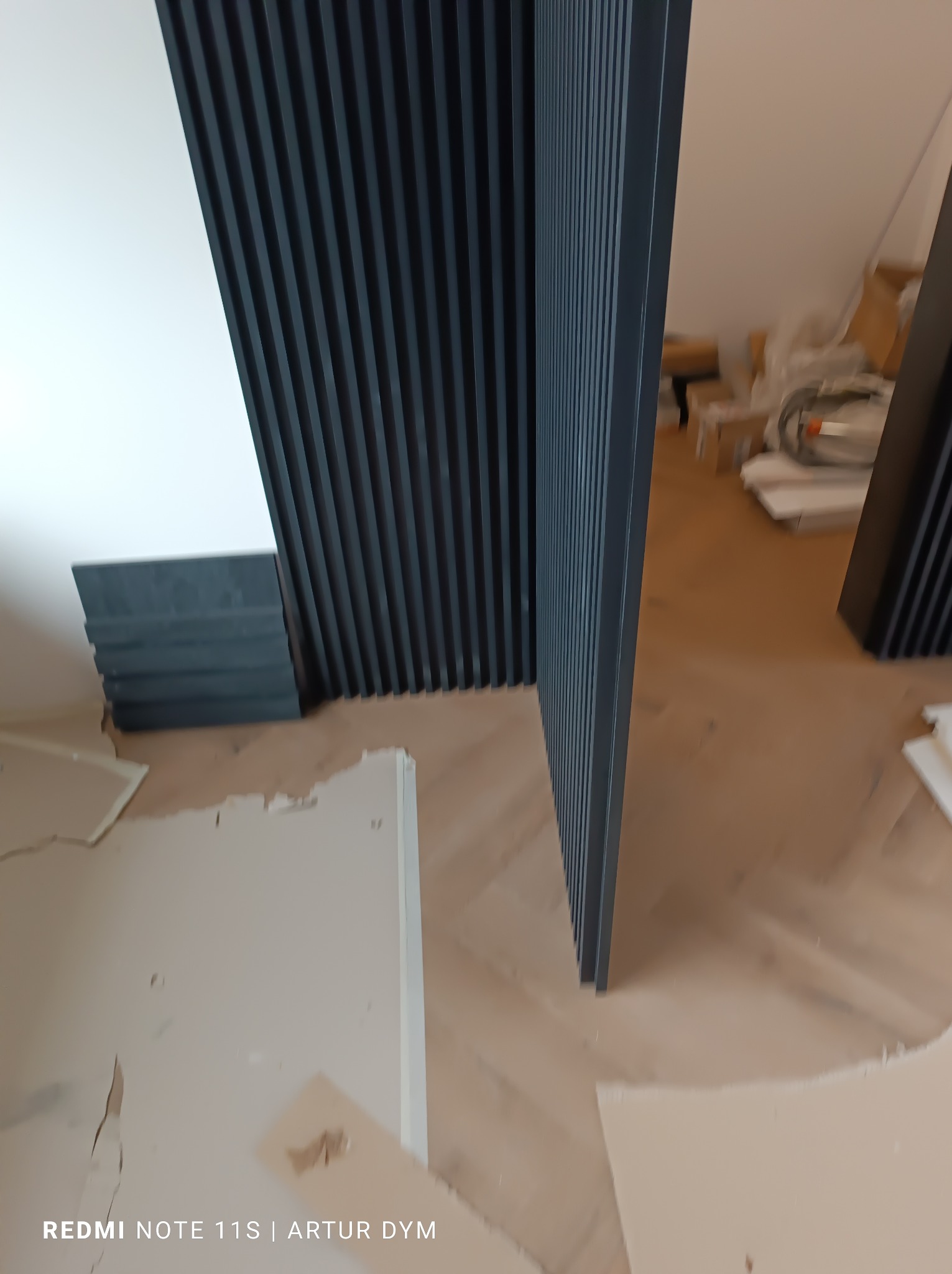 Częściowo zamontowane lamele ścienne w kolorze grafitowym, widoczne niedoskonałości na ścianie i podłodze, wnętrze w trakcie wykończenia.