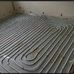 Rozłożona instalacja ogrzewania podłogowego z rurkami przytwierdzonymi do folii izolacyjnej na podłodze w trakcie montażu, widoczne mocowania i oznaczenia na folii.