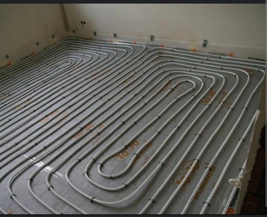 Rozłożona instalacja ogrzewania podłogowego z rurkami przytwierdzonymi do folii izolacyjnej na podłodze w trakcie montażu, widoczne mocowania i oznaczenia na folii.