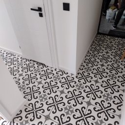 Kacper Podobas - Wzorzyste, czarno-białe płytki podłogowe w przedpokoju, ułożone symetrycznie. Widoczna biała ściana z czarnym włącznikiem i fragment białych drzwi z czarną klamką.
