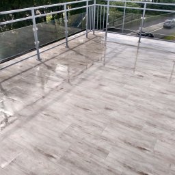 Zdzisław Hamerski - Świeżo ułożona, jasna, drewnopodobna terakota na balkonie z metalową balustradą i szklanymi panelami, mokra od deszczu, odbijająca światło.