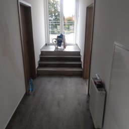 Zdzisław Hamerski - Korytarz w trakcie prac wykończeniowych z szarymi panelami podłogowymi imitującymi drewno, białe ściany, grzejnik, butelka wody i urządzenie do malowania natryskowego na schodach prowadzących do okna.